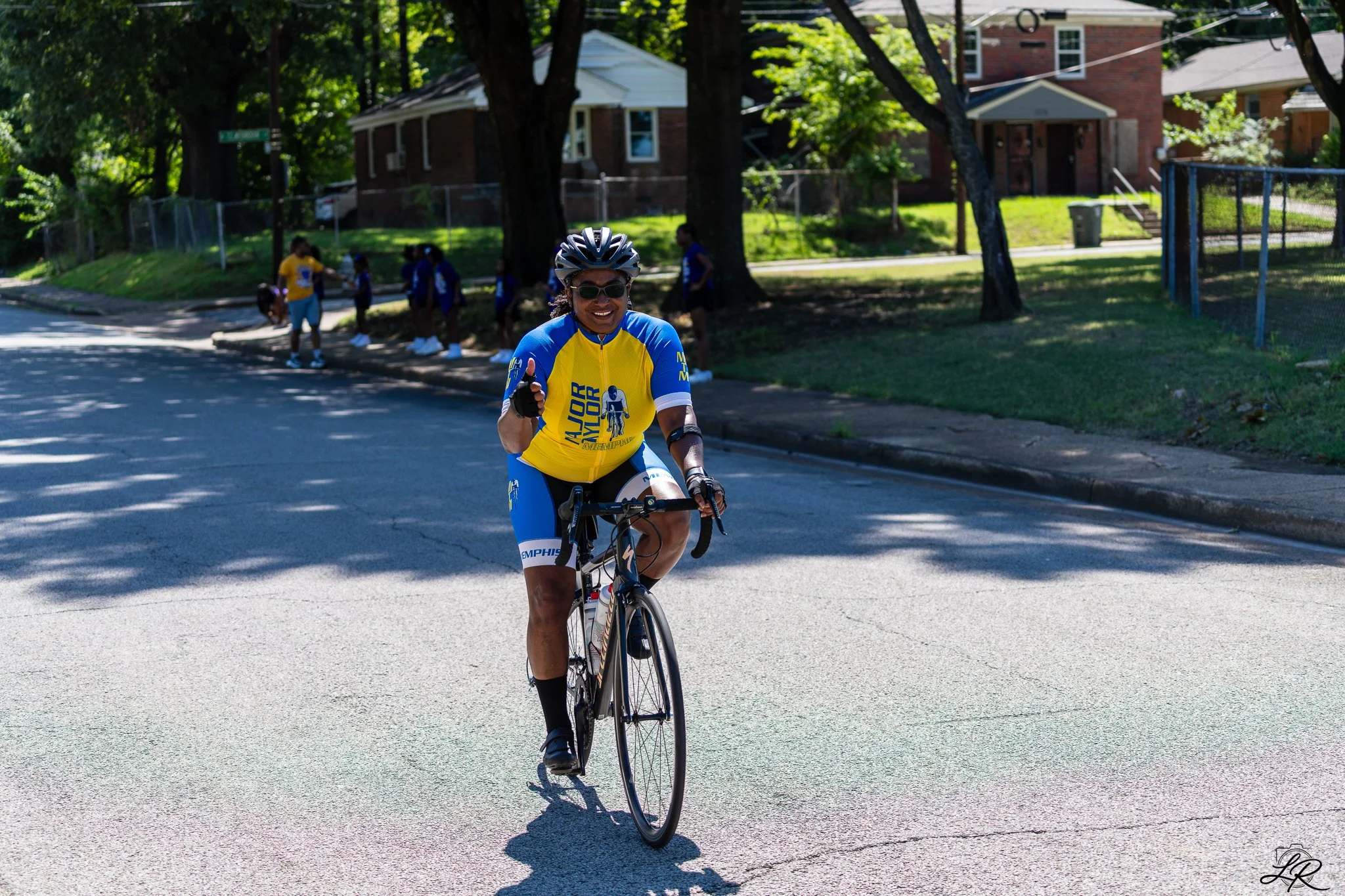 A woman riding a bicycle on a quiet street, wearing a helmet and sunglasses, with a group of children in the background near houses and trees.