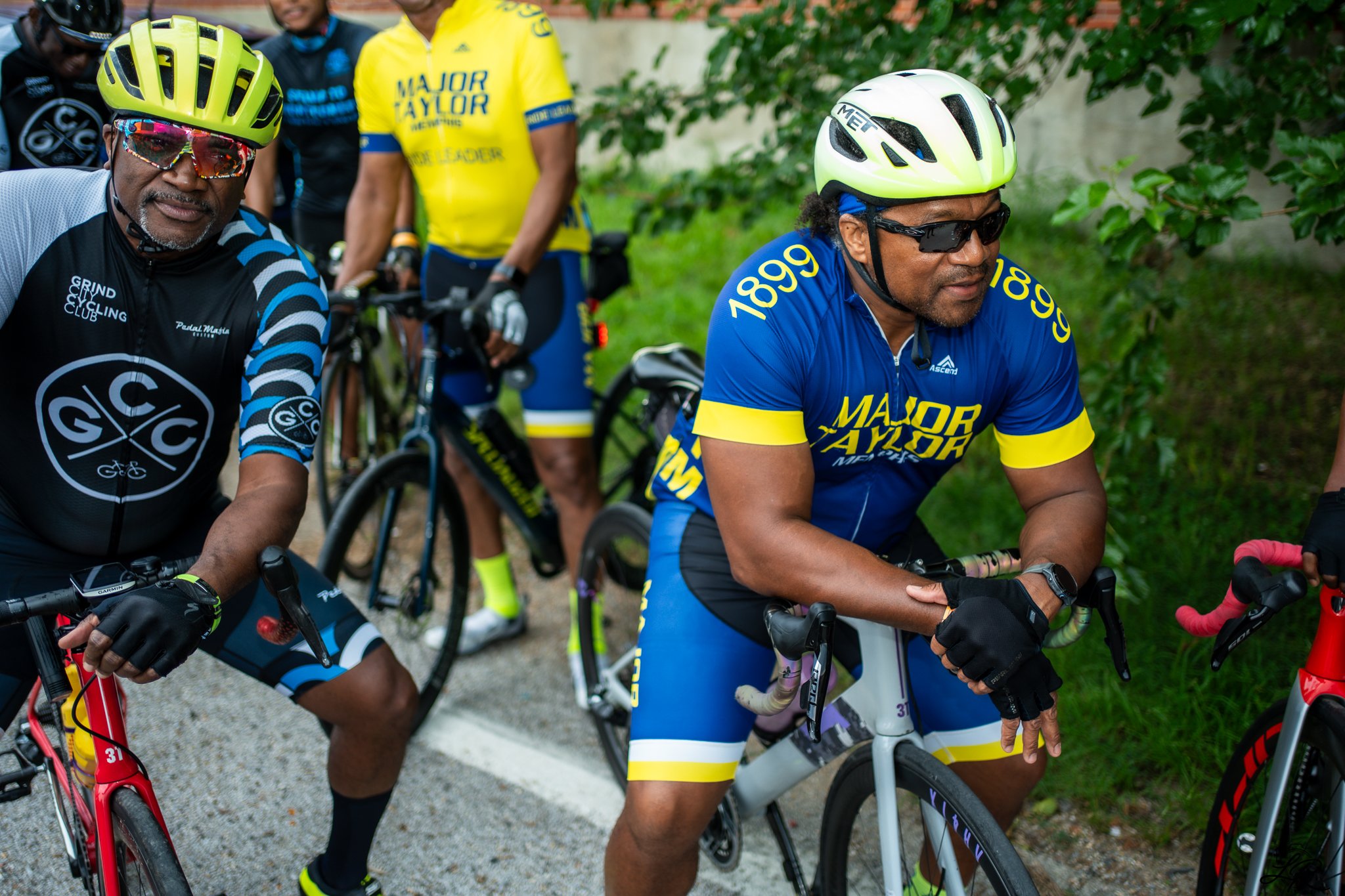 A group of people dressed in cycling gear, some with helmets and sunglasses, standing with their bicycles outdoors near greenery.