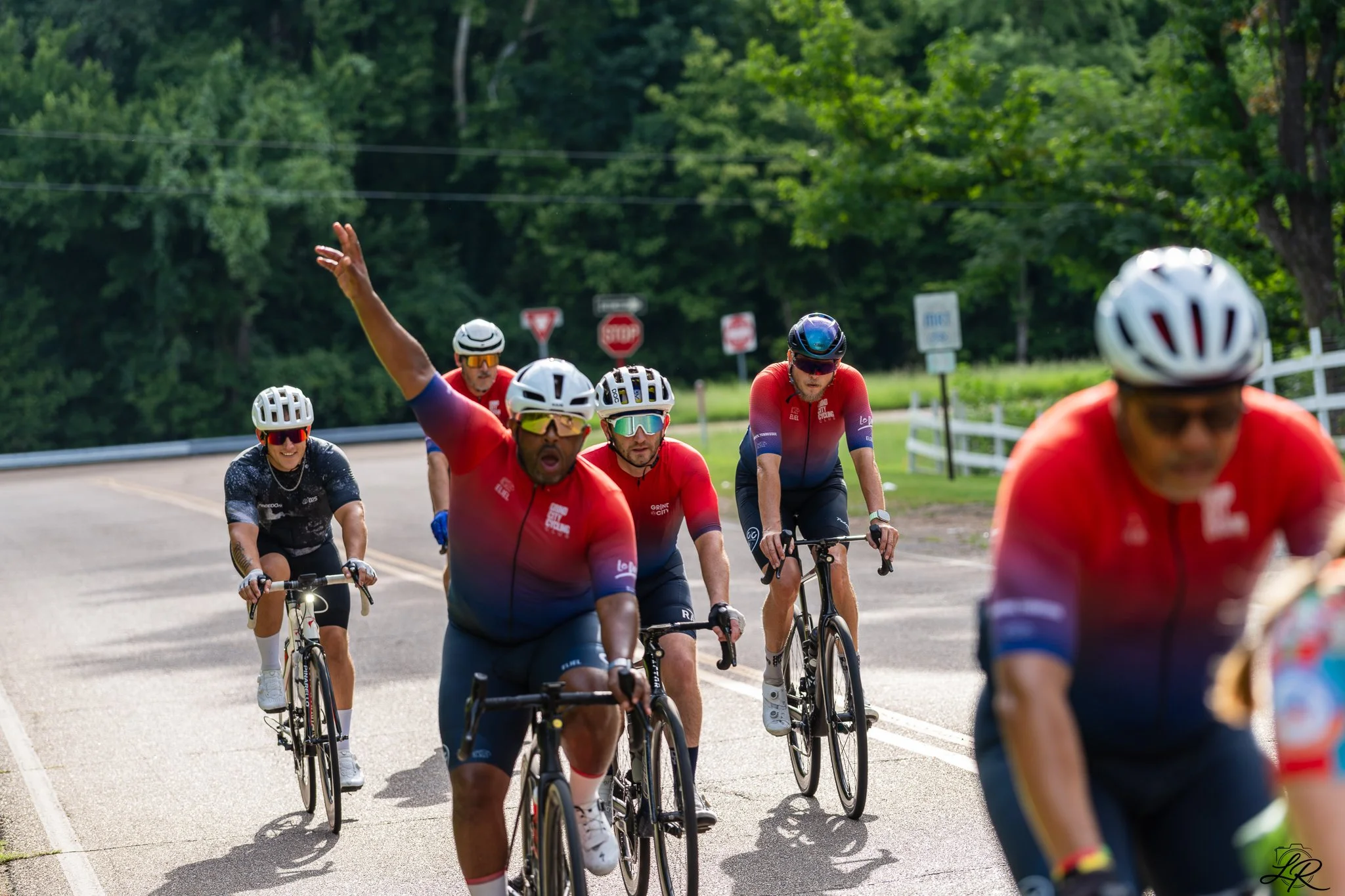 Group of cyclists wearing helmets riding on a tree-lined road during daytime, with a mostly sunny sky.