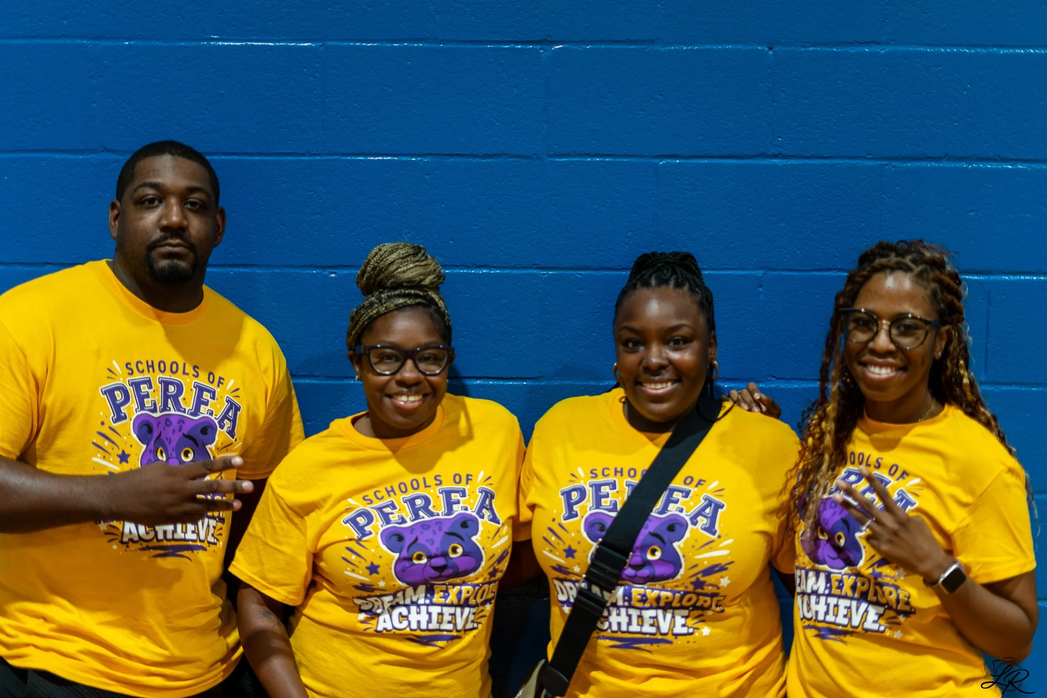 Group of five people wearing yellow T-shirts with a purple bear graphic and the words 'Schools of PERA' and 'Team: Explore, Achieve, Believe' written on them, standing against a blue wall.
