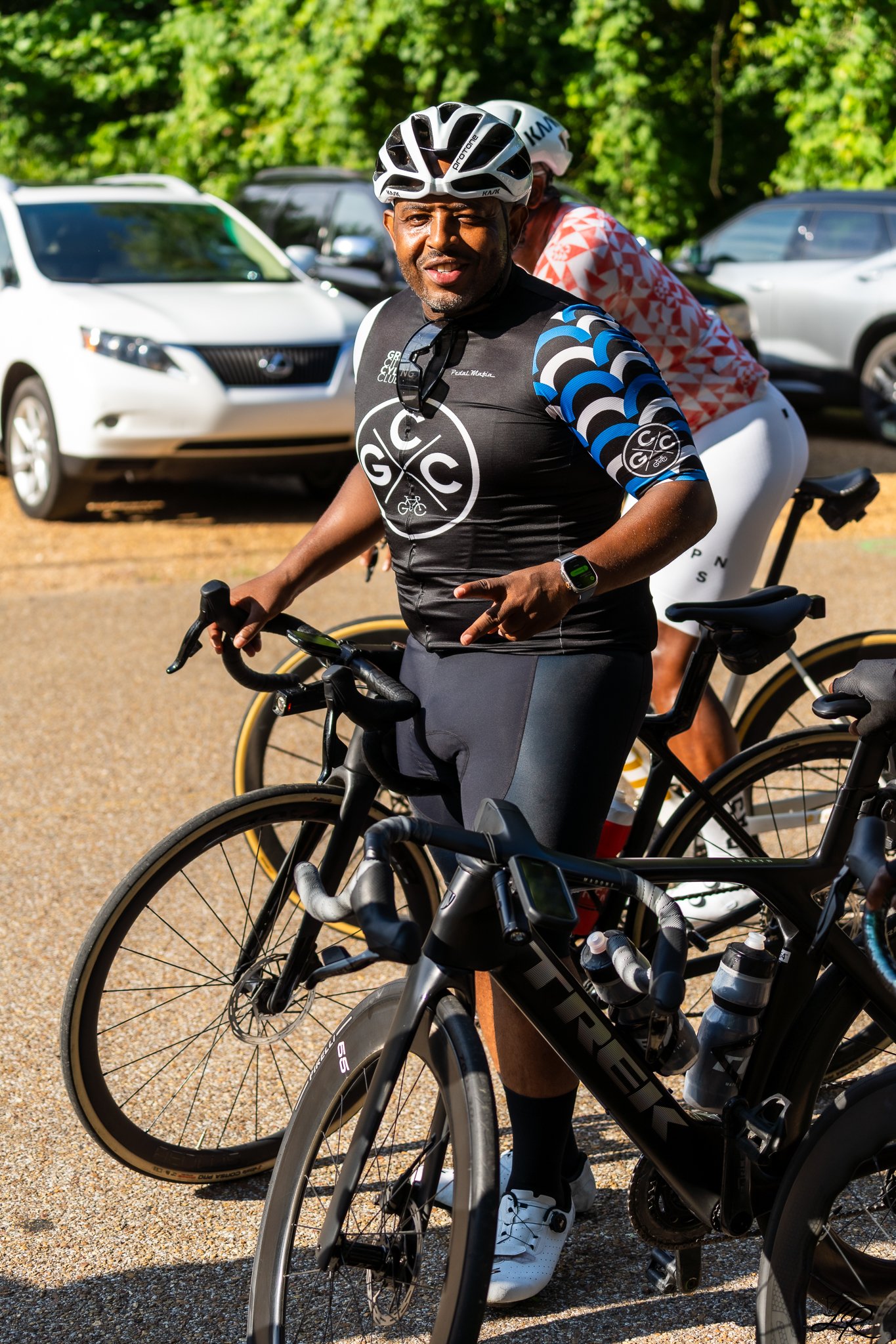 Man in cycling gear and helmet riding a bicycle, with another cyclist behind him, surrounded by parked cars and green trees.