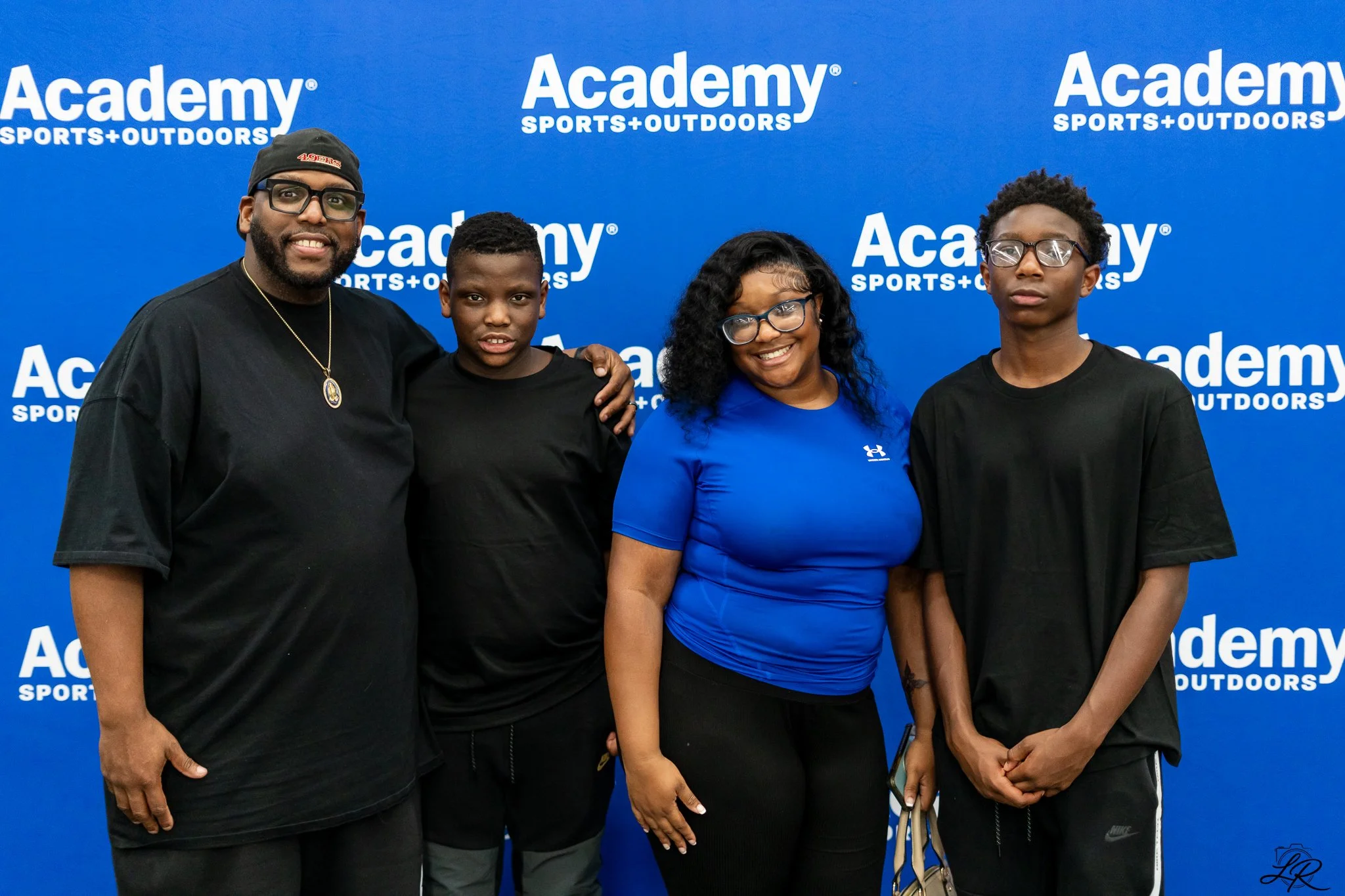 Group of four people standing in front of a blue backdrop with 'Academy Sports + Outdoors' logo, posing for a photo.
