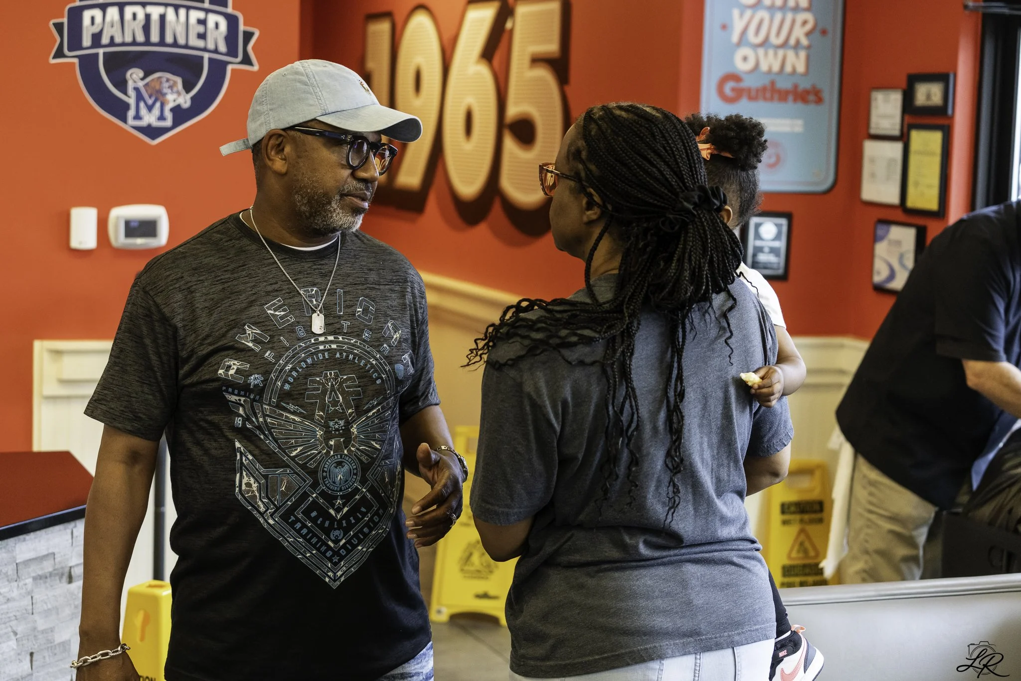 Two people are talking inside a restaurant or cafe with red walls and various signs, including a large "106.5" in the background. One person is wearing a gray cap, glasses, a graphic black T-shirt, and jewelry, while the other is wearing glasses, a g