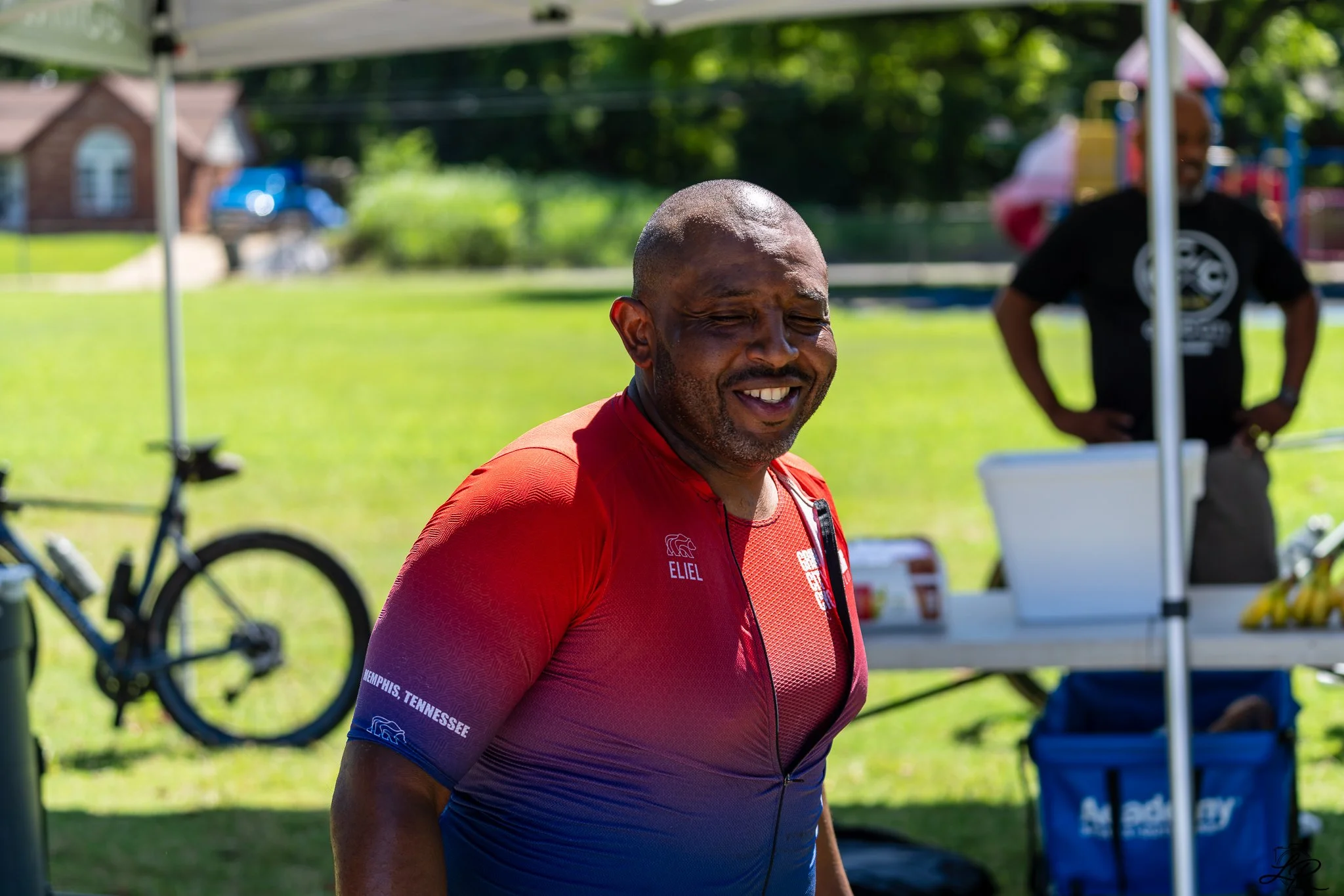 A man in a red and blue athletic shirt standing outdoors with a smile, with a woman in a black shirt and colorful hat in the background at a park or outdoor event.