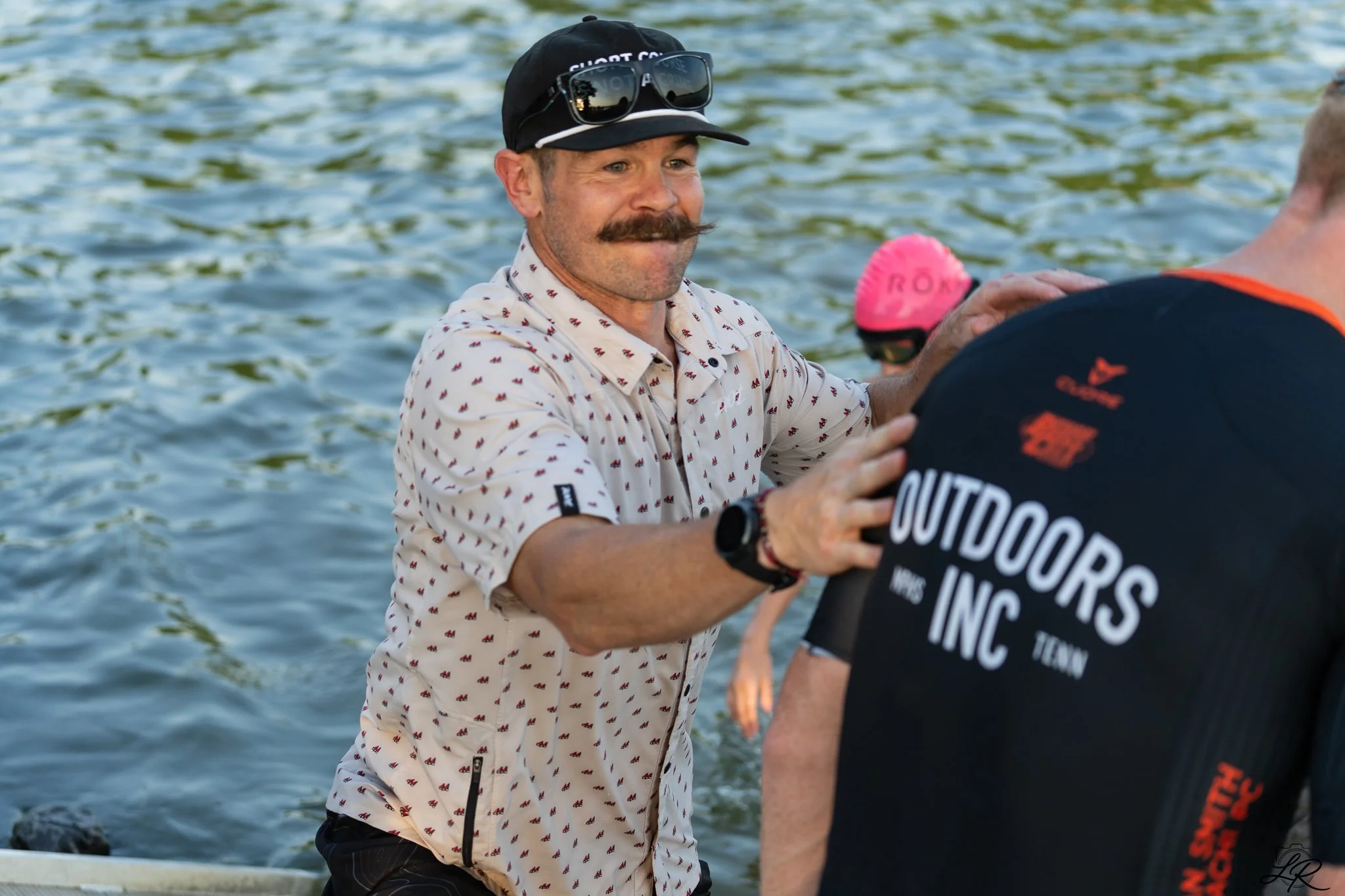 A man with a mustache wearing sunglasses, a black cap, and a printed button-up shirt, standing in a body of water, holding hands with another person whose back is visible, wearing a black T-shirt with orange and white text.