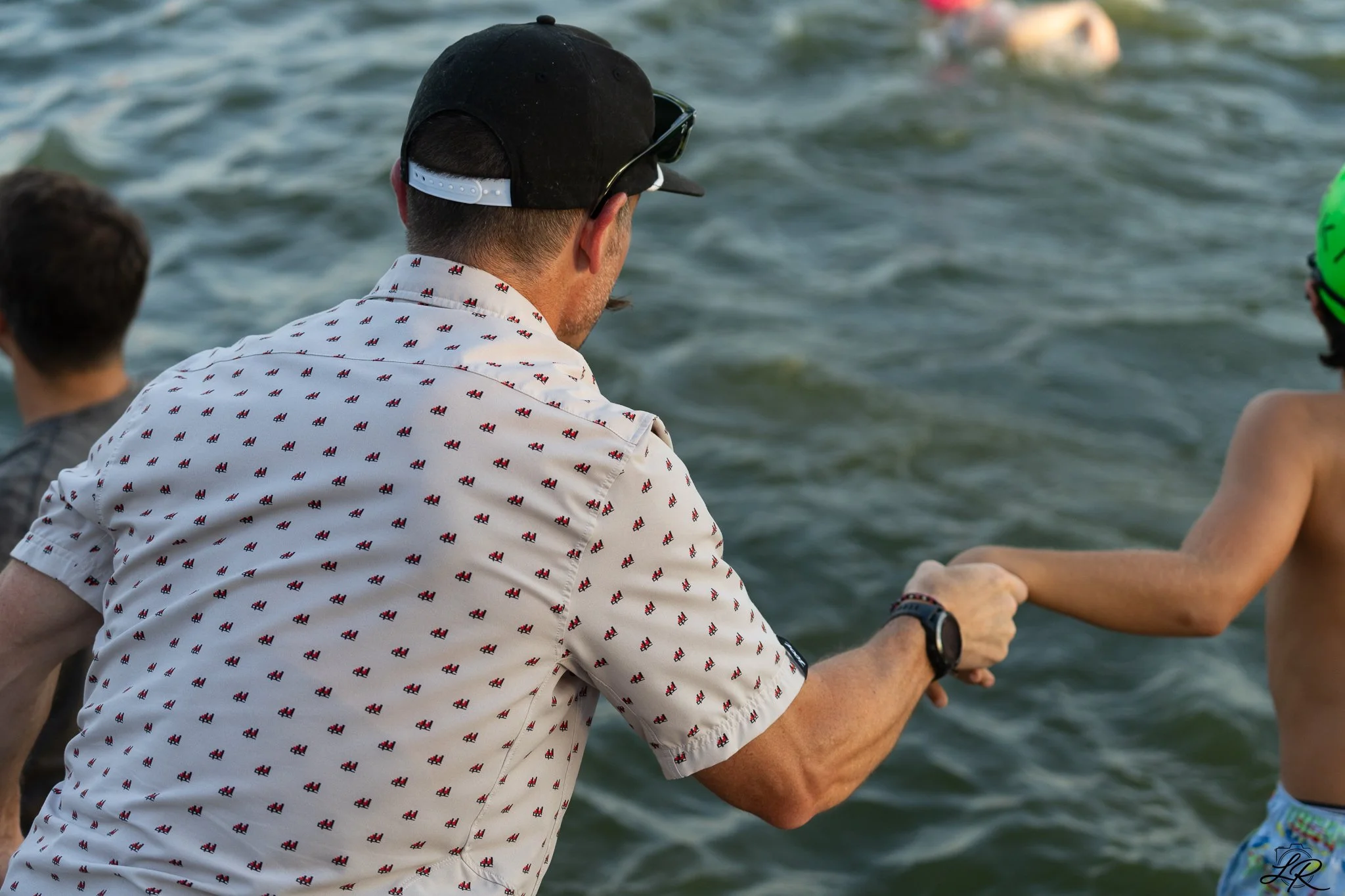 A man wearing a black baseball cap, sunglasses, a white shirt with small red and blue pattern, and a wristwatch, sitting near water, holding the hand of a person with a colorful bracelet. In the background, a person with a green helmet is visible