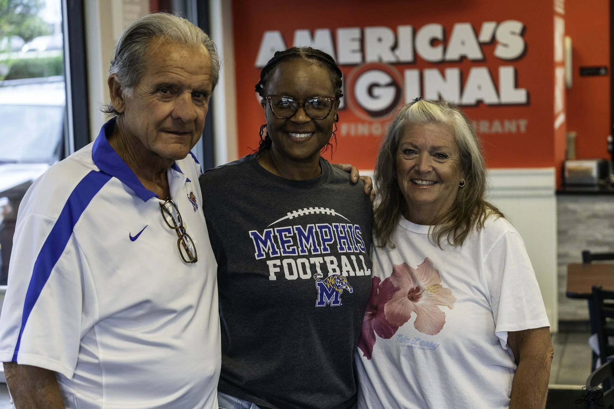 Three women standing together inside an office with a Memphis Football shirt worn by the middle woman, a large red sign in the background that says Americansignal, and large windows on the side.