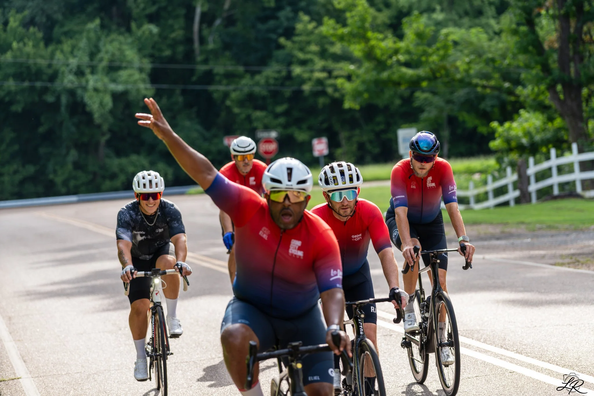 A group of cyclists riding on a sunny, tree-lined road, with one cyclist in the front raising his arm in greeting or celebration.