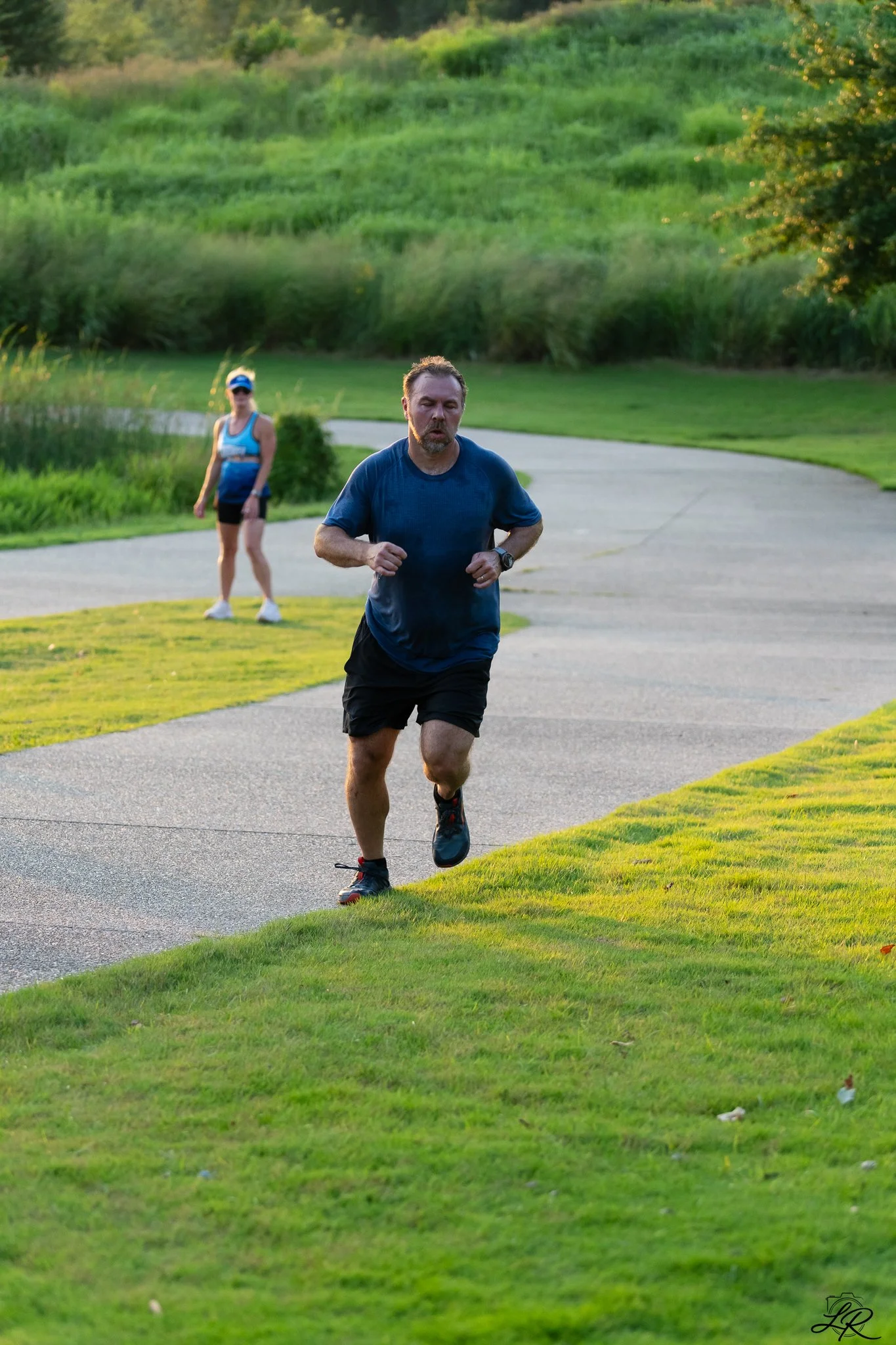 A man jogging along a paved park pathway with a woman walking behind him, lush green grass and trees in the background during the evening.