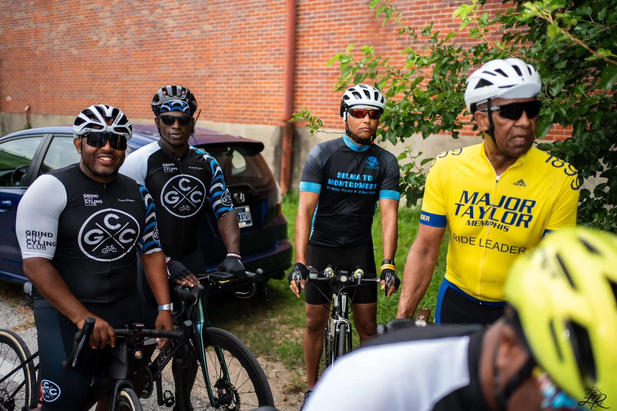 Group of four cyclists in helmets and sunglasses standing outdoors near a brick wall and greenery.