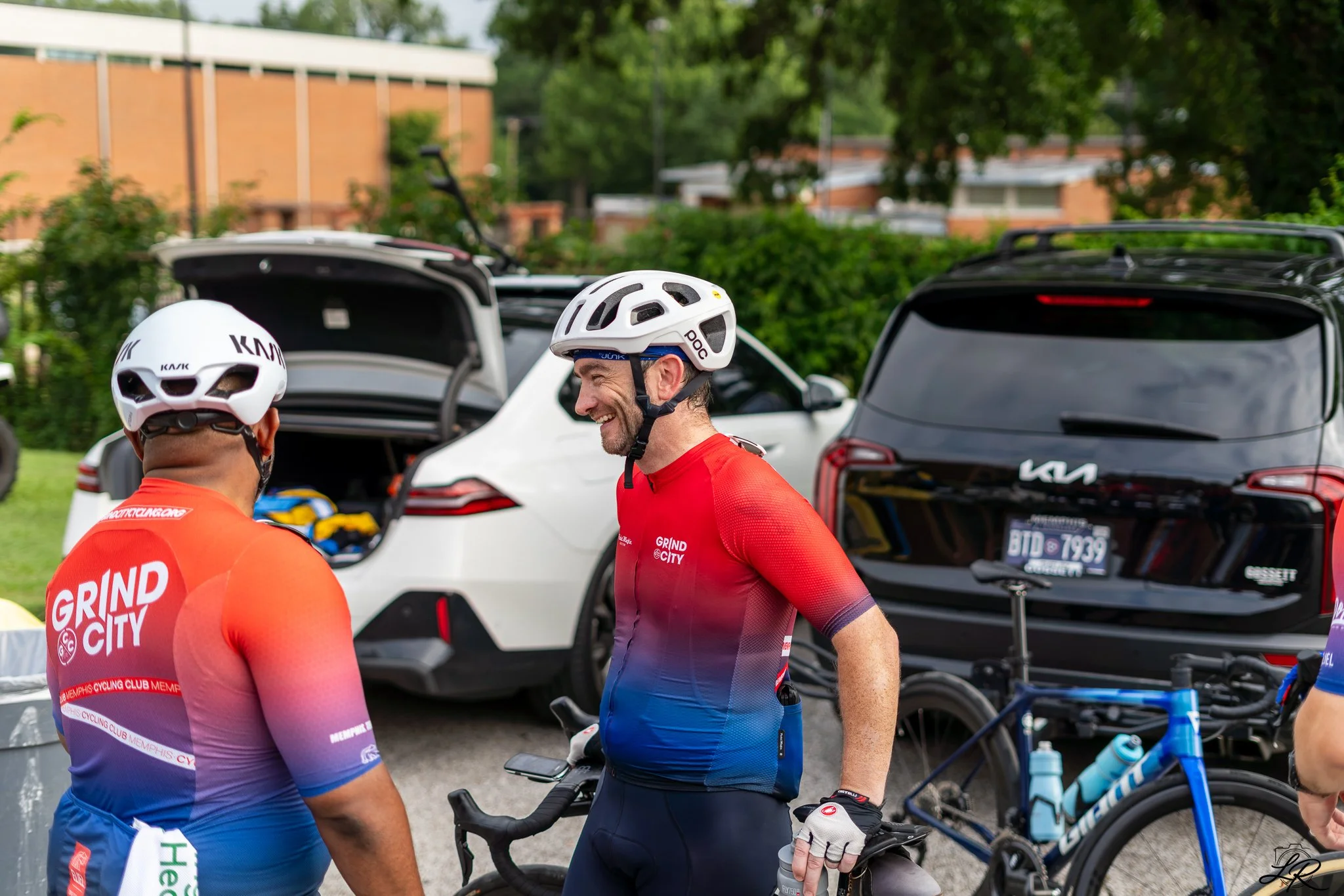 Two men in cycling gear and helmets, smiling and talking next to parked cars and bicycles in an outdoor setting.