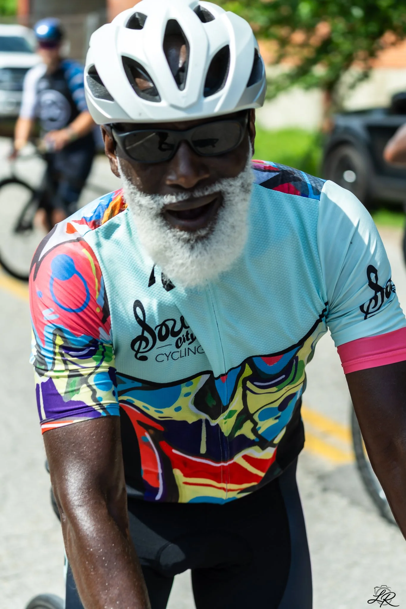 Close-up of an elderly man with a white beard, wearing a colorful cycling jersey, black sunglasses, and a white bike helmet, riding a bicycle outdoors.