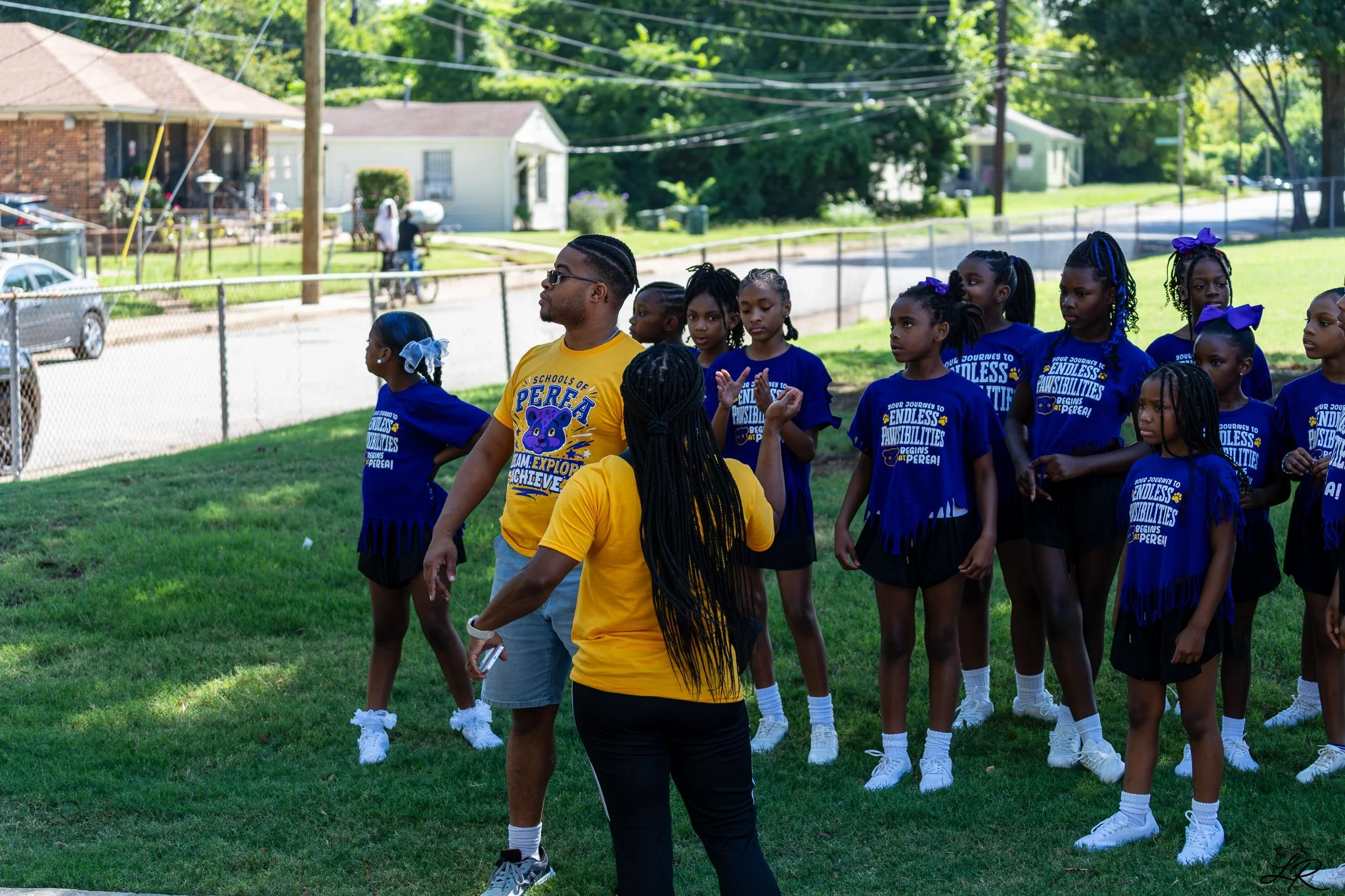 A group of young girls in blue cheerleading uniforms with purple bows standing outdoors on a grassy field, with two adult women walking among them, one wearing a yellow shirt and the other in a black shirt. The background shows residential houses, tr