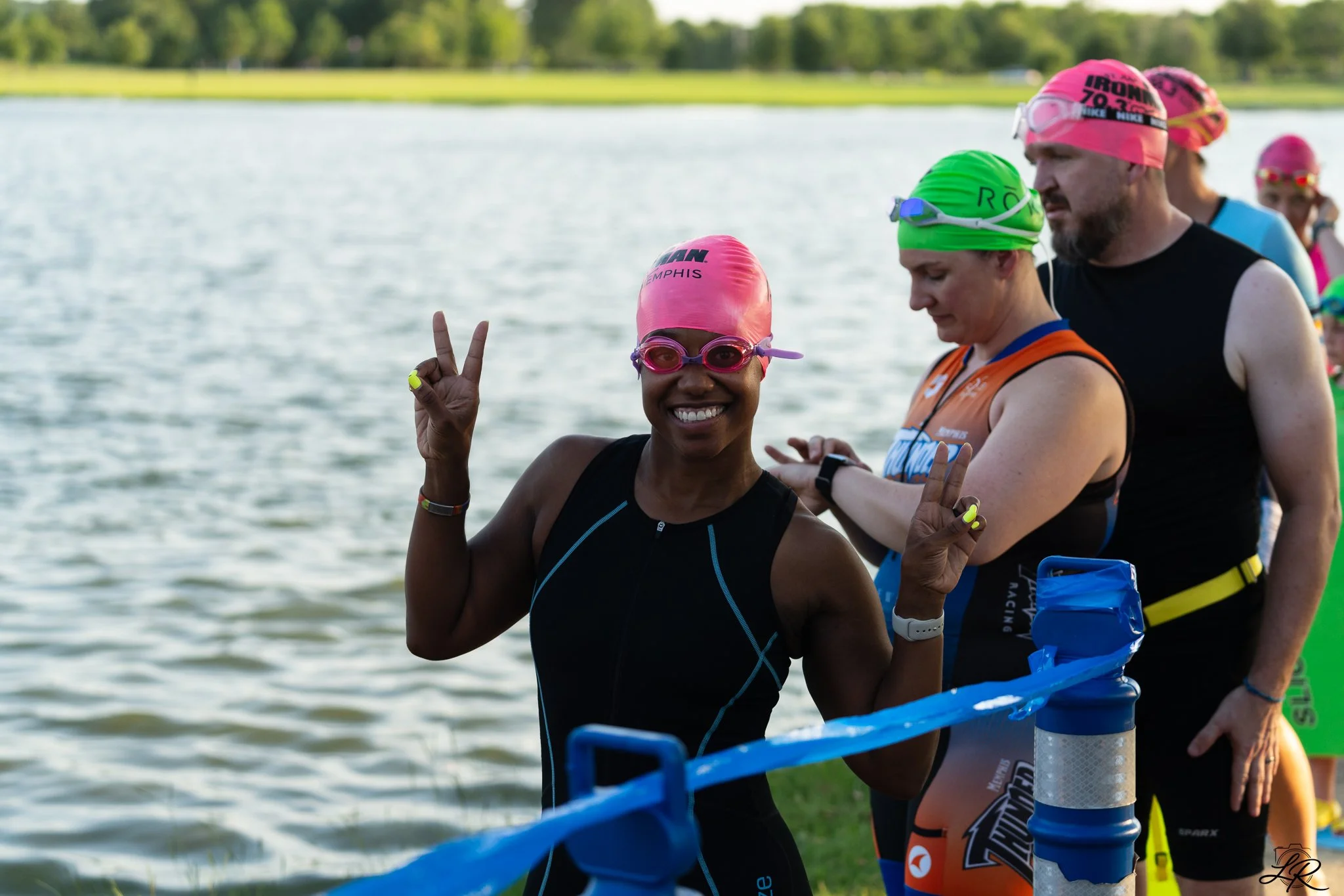 Group of people dressed in colorful triathlon gear, standing by water, preparing for a race.
