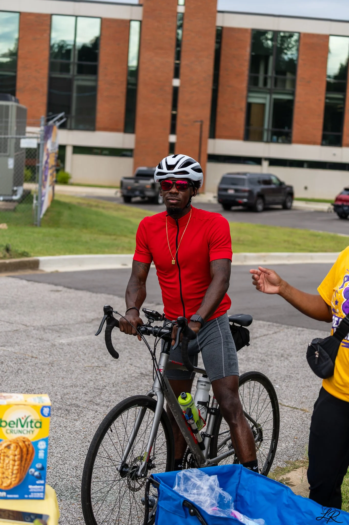 A man in a red shirt and black cycling shorts wearing a helmet and red sunglasses is standing with his bike outdoors in front of a brick building. There is a blue trash bin and food packages nearby.