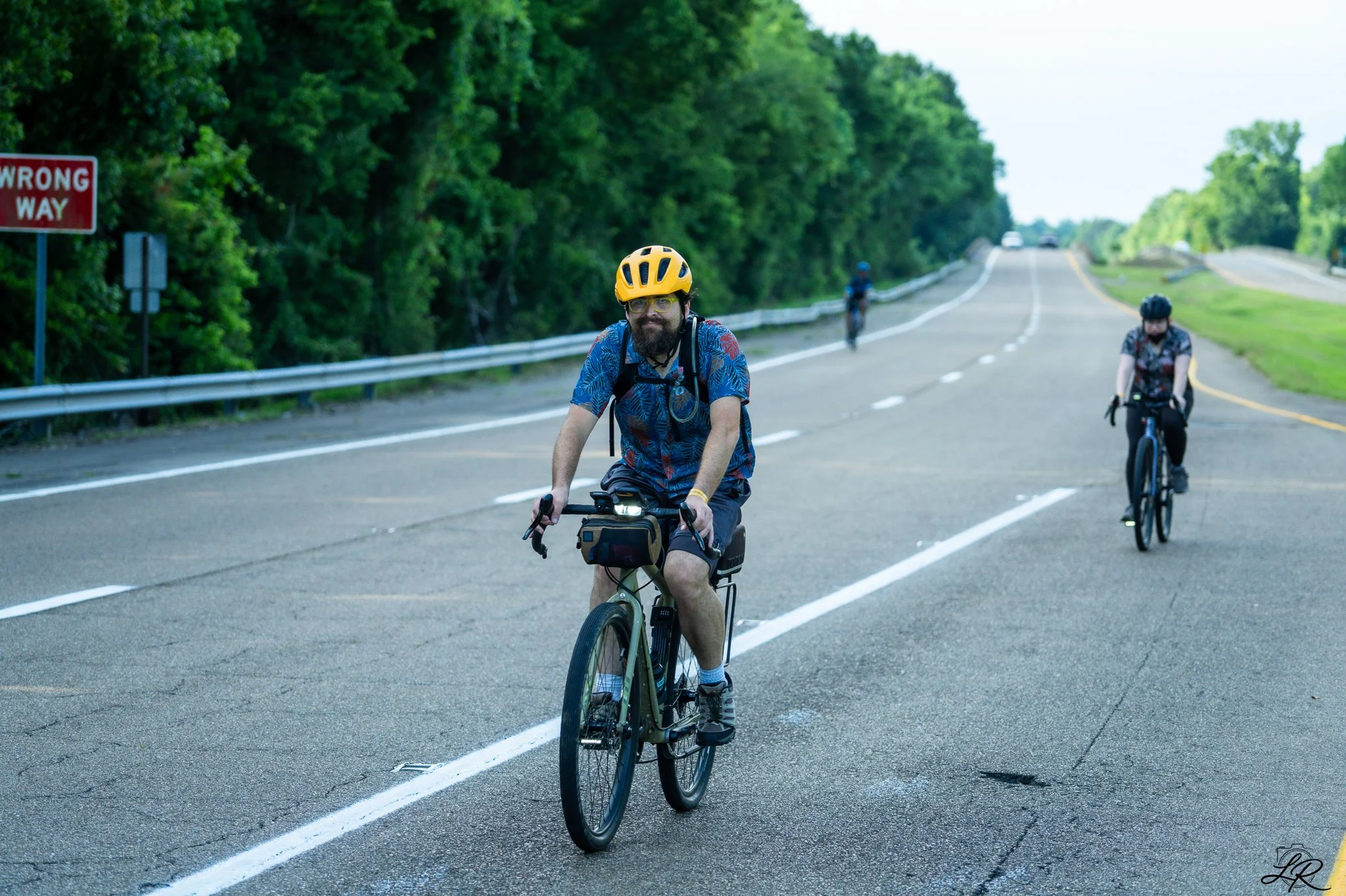 Cyclists riding on the side of a rural road surrounded by green trees, with a red 'Wrong Way' sign on the left.