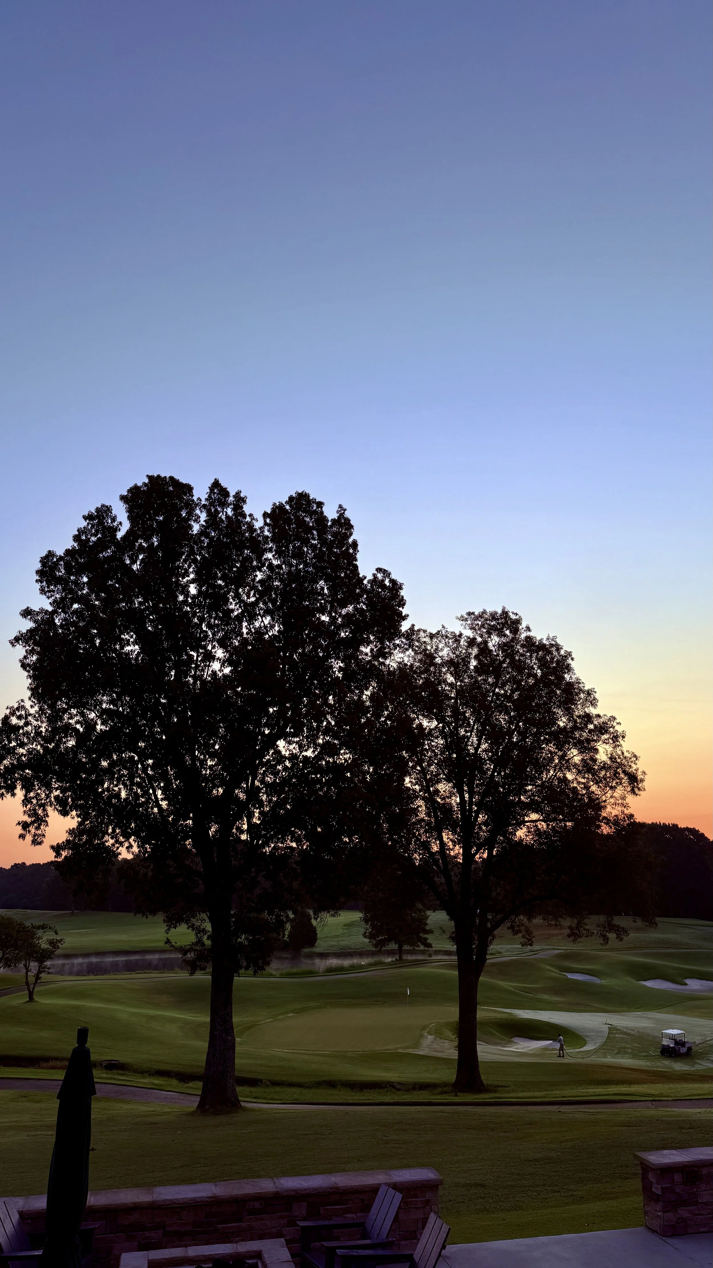 A golf course at sunset with two large trees in the foreground, green fairways, sand bunkers, and a person walking near the sand traps, all under a twilight sky.