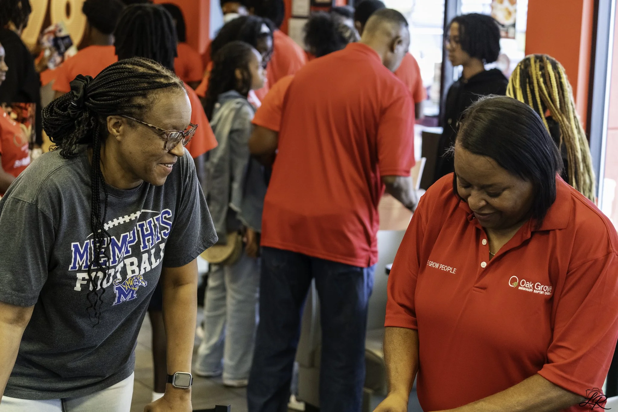 A group of people standing in line at a restaurant or fast food place, waiting to order. Two women in the foreground are engaged and smiling, one wearing a Memphis football shirt and the other wearing a red Oak Grove shirt.