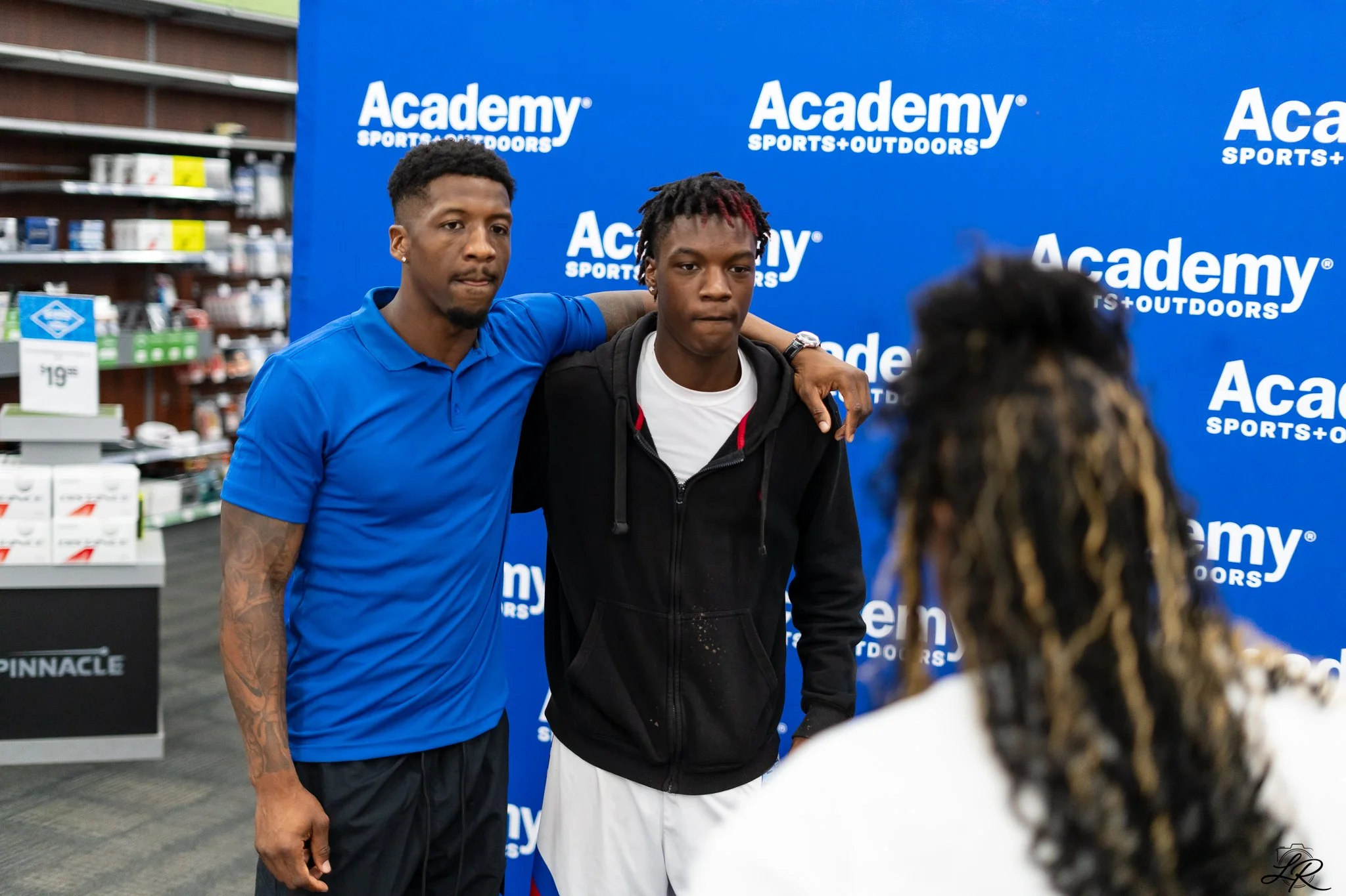 Two young men pose together with arms around each other in front of a blue backdrop with 'Academy Sports + Outdoors' logo. One wears a blue polo shirt, and the other wears a black hoodie and white shorts, inside a retail store.