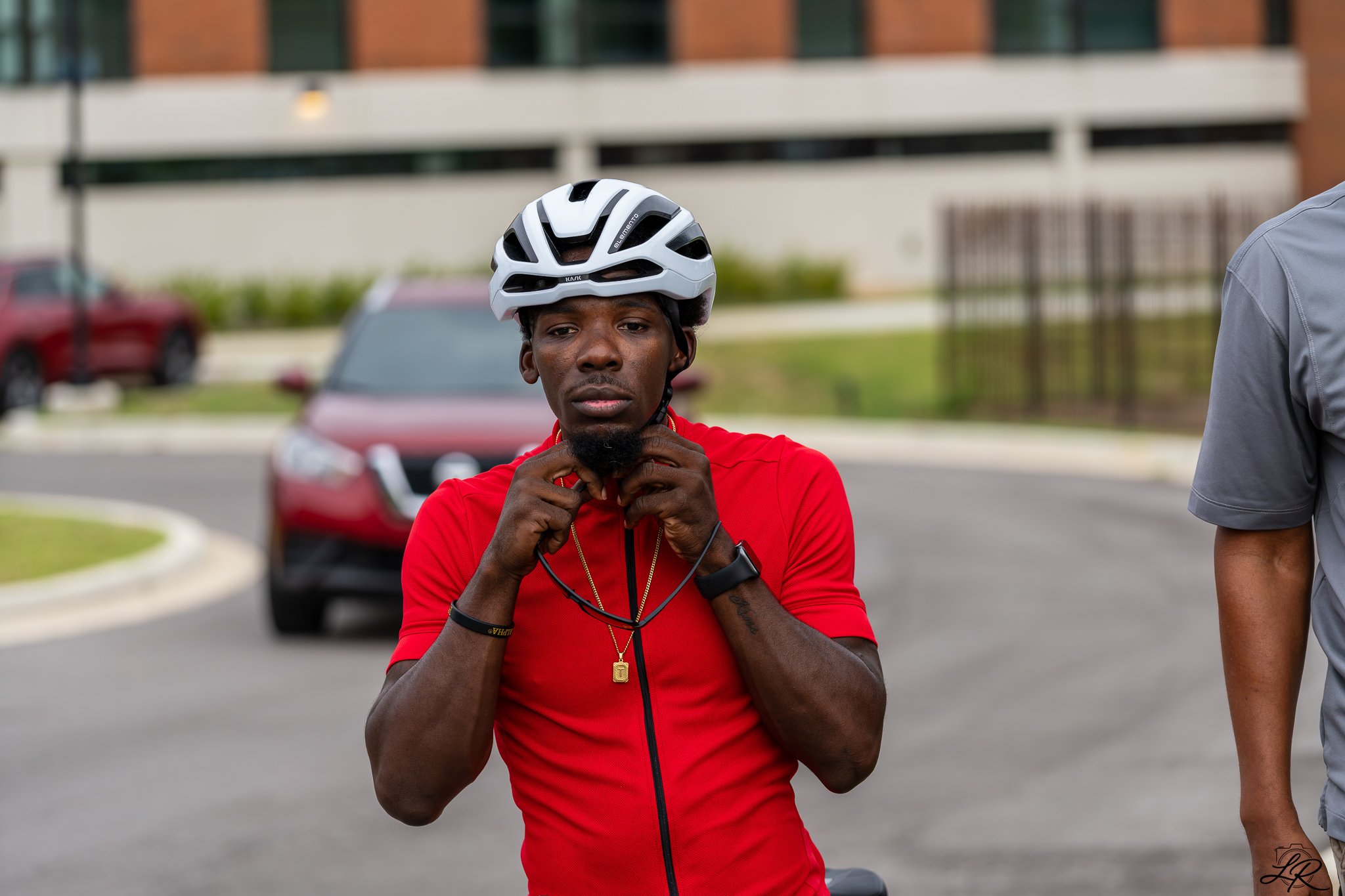 A young man with dark skin and short hair wearing a white bicycle helmet, red shirt, and gold chain, adjusting his helmet strap outdoors near parked cars and a residential building.