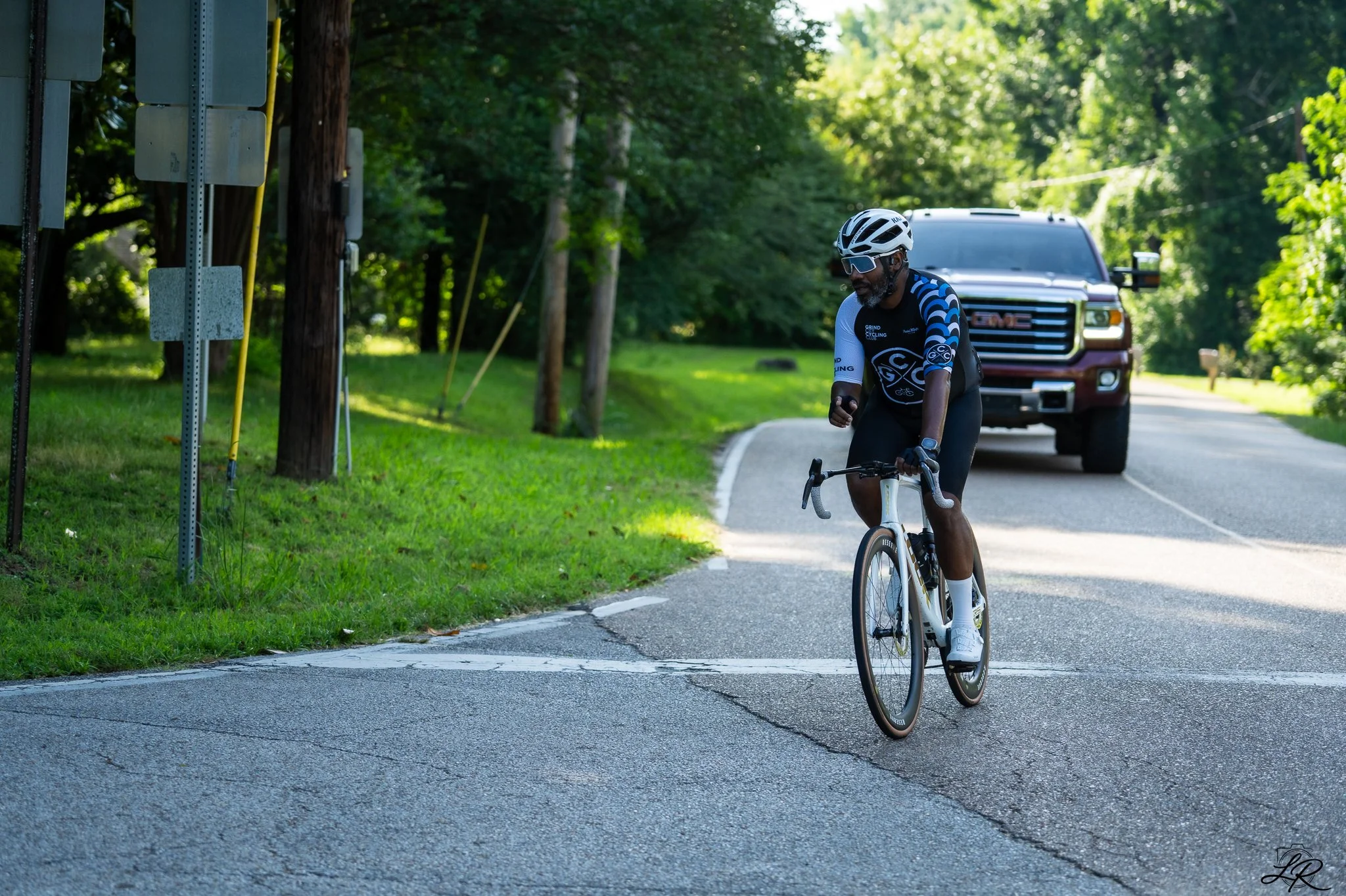 A man wearing a helmet, sunglasses, and cycling attire riding a bicycle on a suburban road with trees and a pickup truck in the background.