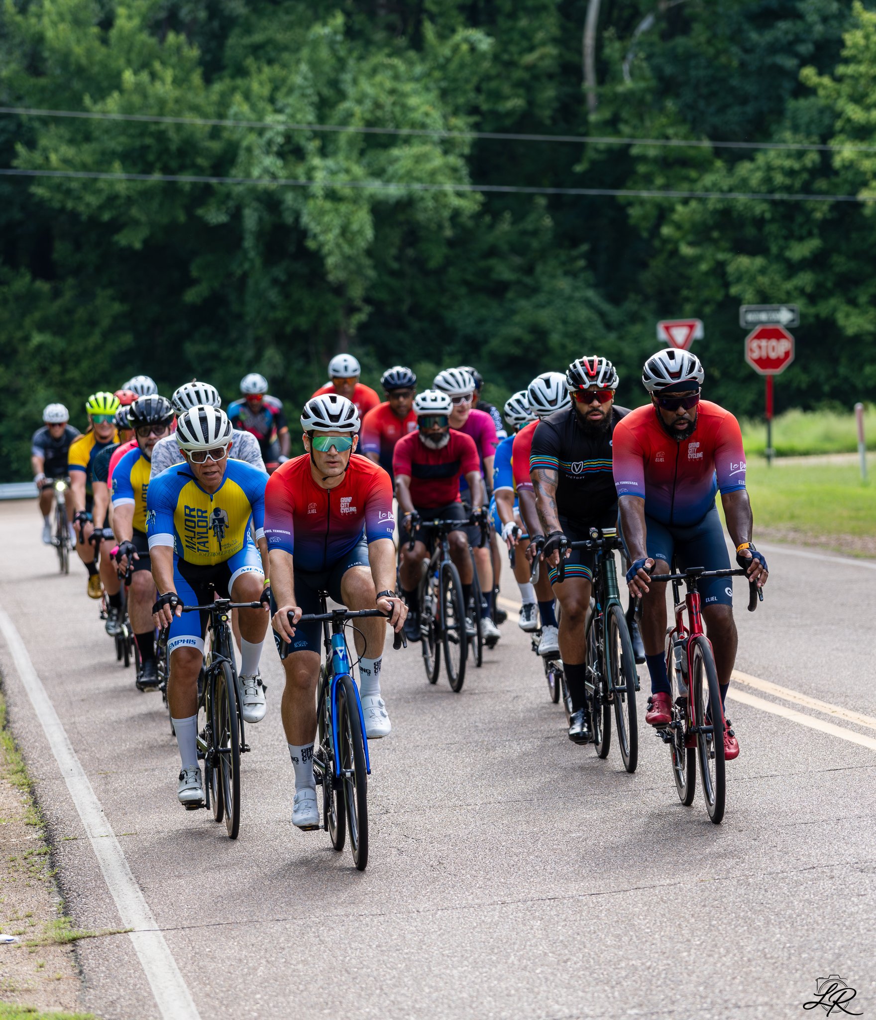 A large group of cyclists riding together on a road, surrounded by trees and greenery.