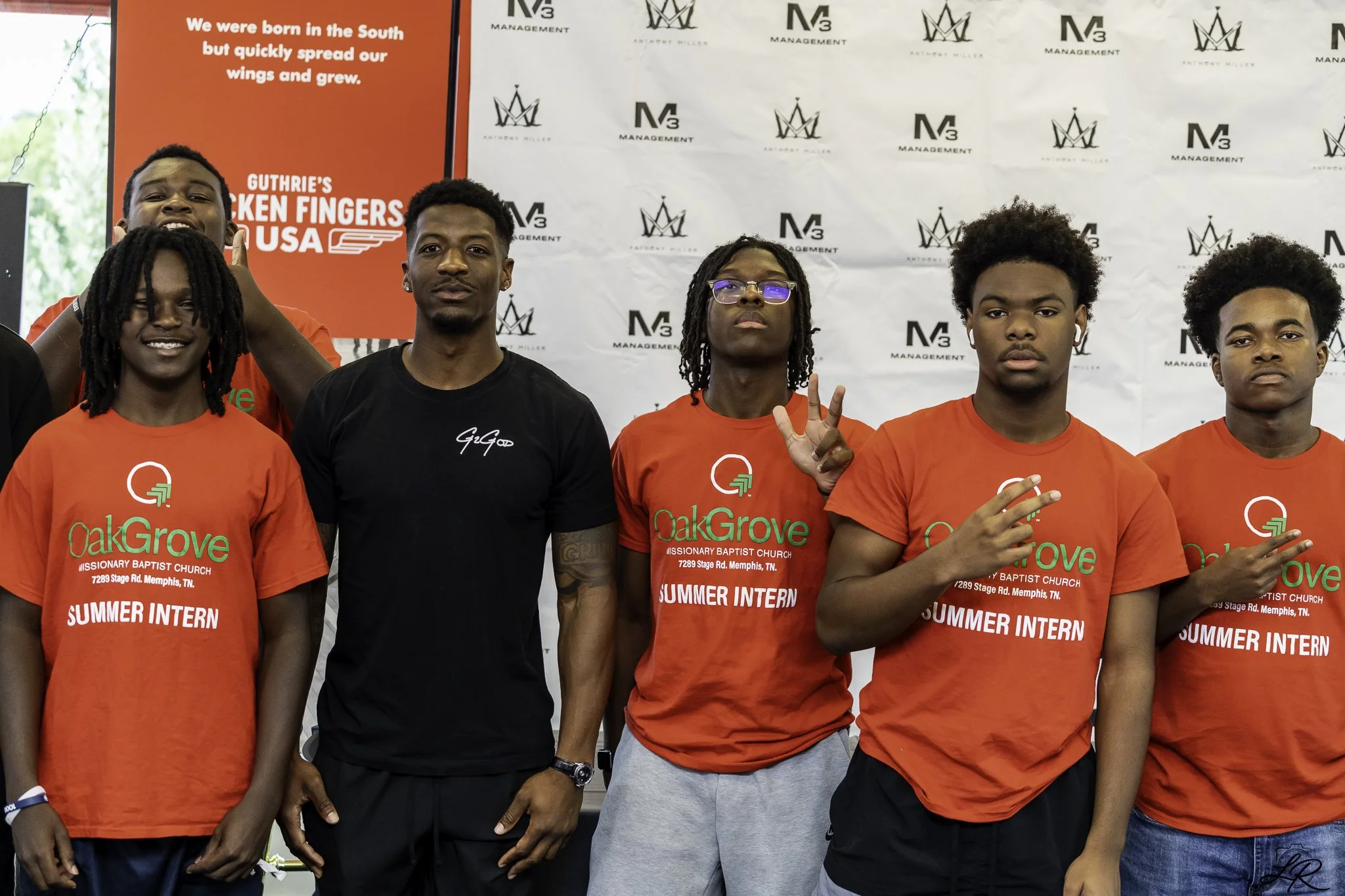 Group of young men and women posing in front of a backdrop with logos, some wearing red shirts that say 'Summer Intern' and 'OakGrove Missionary Baptist Church' and others in casual black clothing. One woman is making a peace sign.