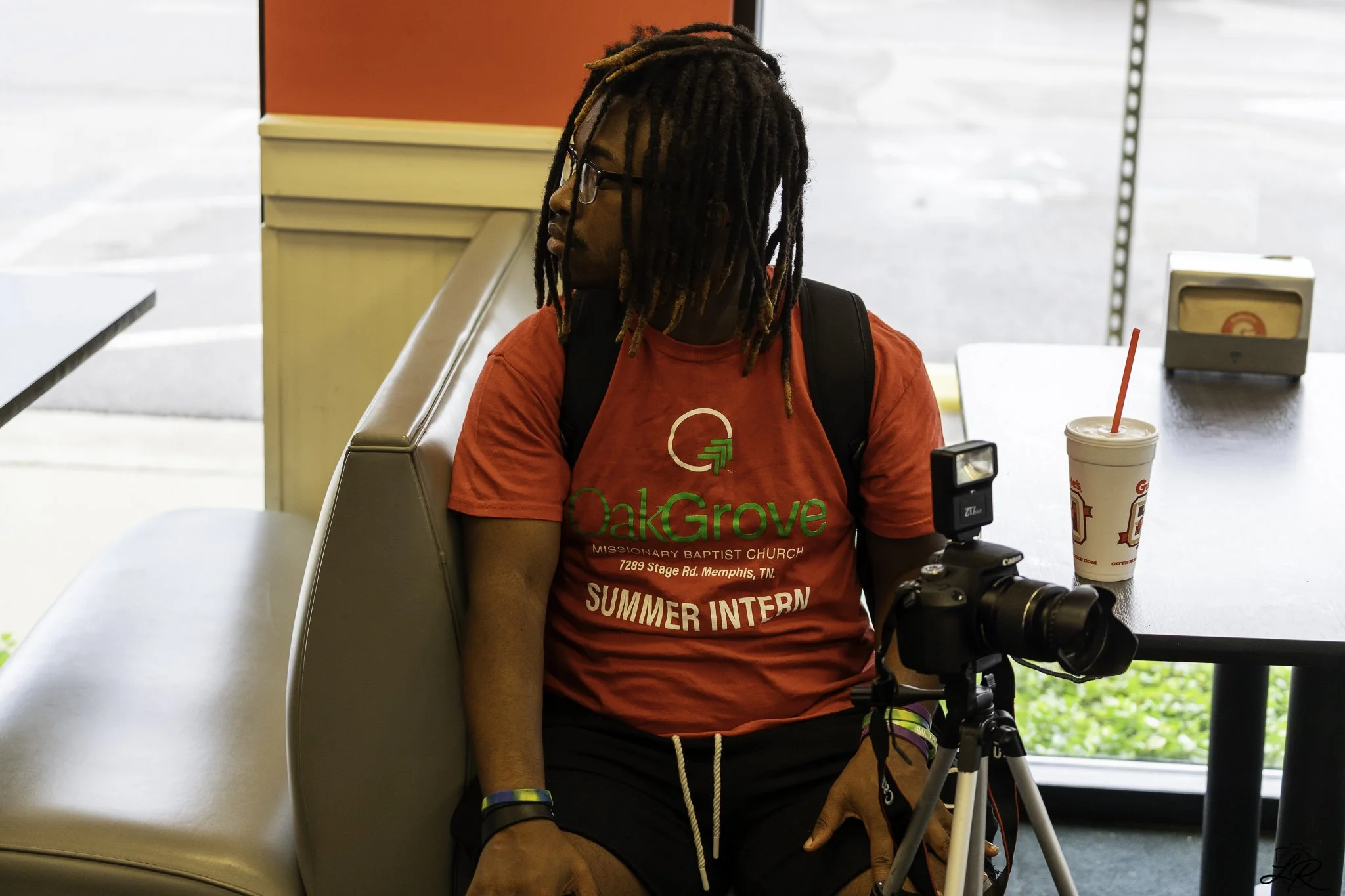 A person with long dreadlocks, glasses, and a red OakGrove Summer Intern t-shirt is sitting at a table in an indoor setting, with a beverage and camera in front of them.