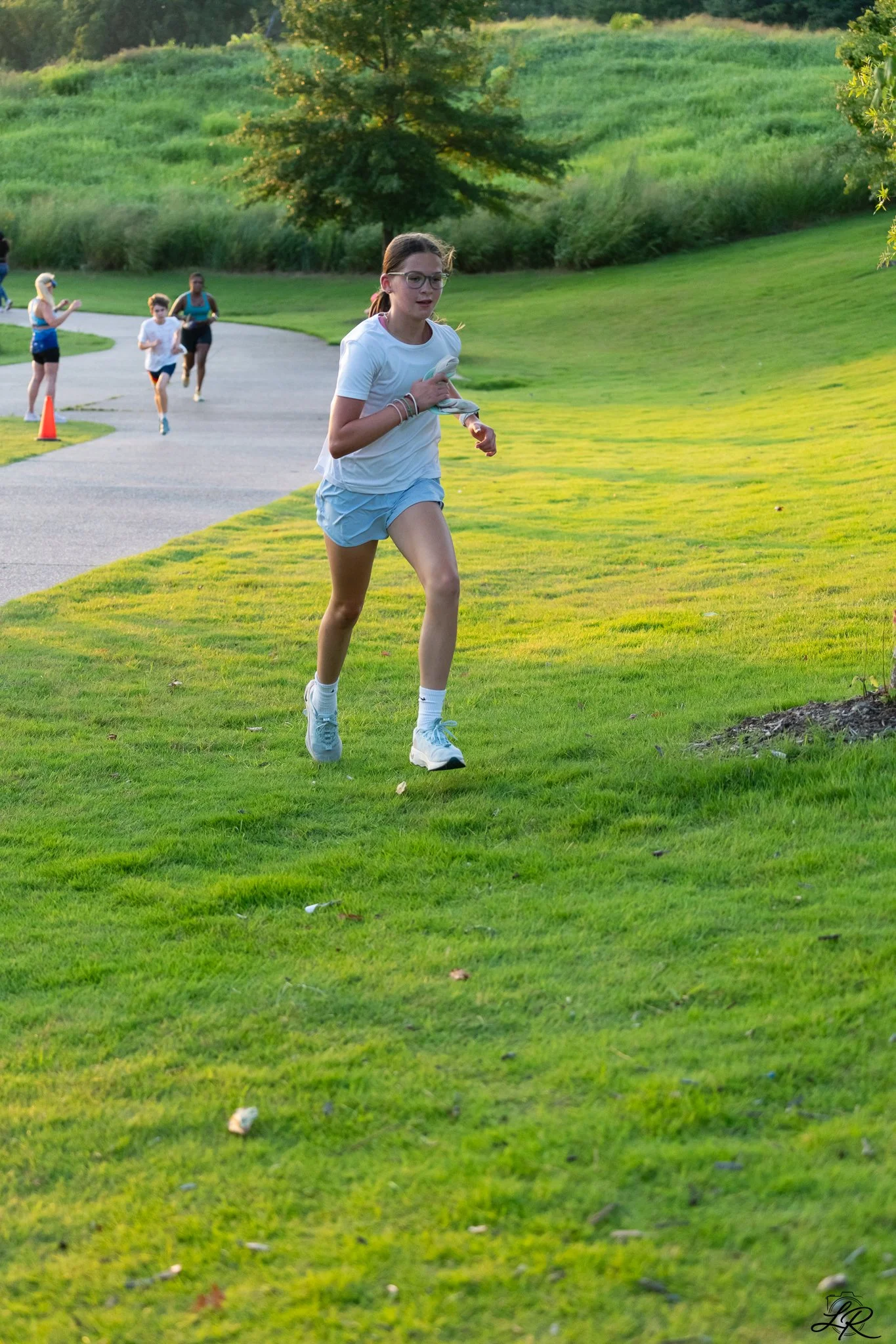 A young girl running outdoors on a grassy area with trees in the background. She has glasses, a white t-shirt, light blue shorts, and white sneakers. Other children are running or standing on a paved path in the background.
