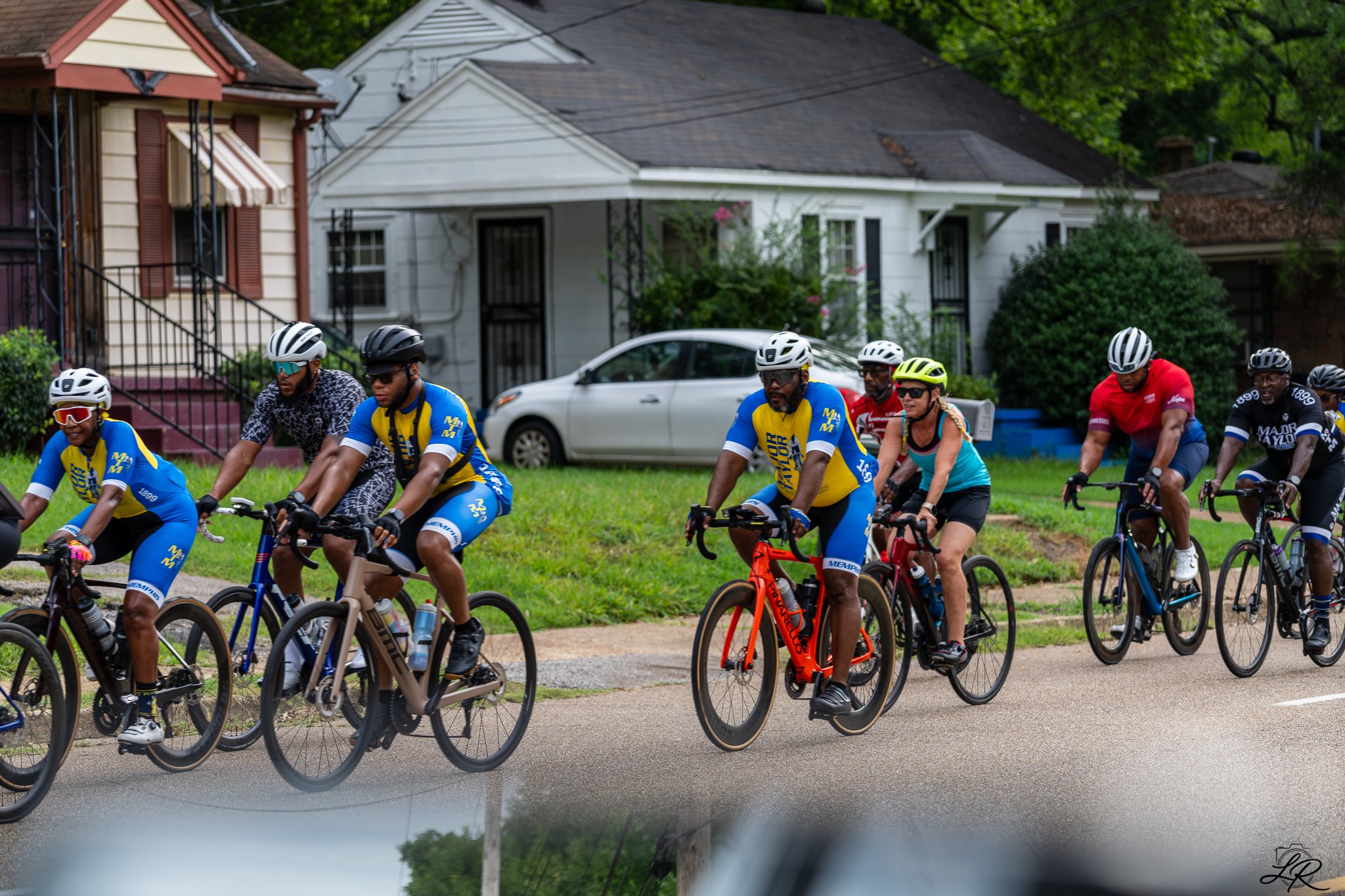 A group of people riding bicycles outdoors in front of houses and trees.