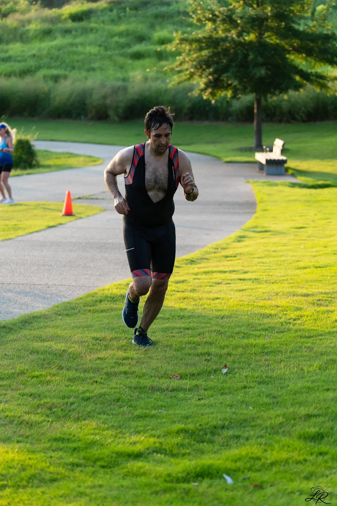 A man running outdoors along a curved paved path surrounded by green grass, trees, and a park bench, during daylight hours.