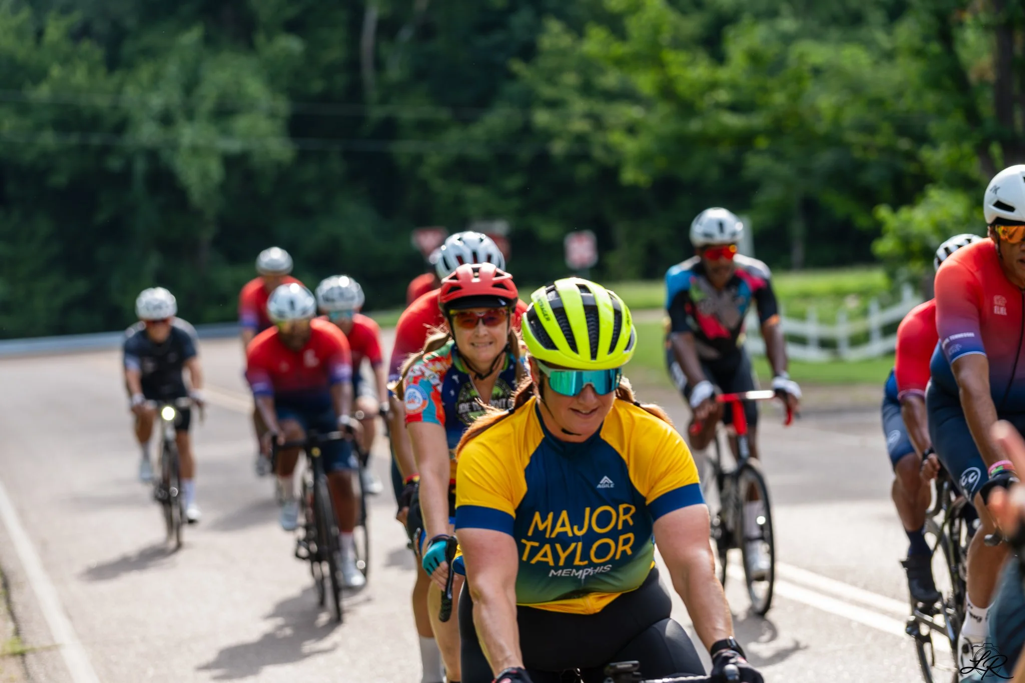 Group of cyclists riding on a road surrounded by greenery, with some wearing helmets and sportswear.
