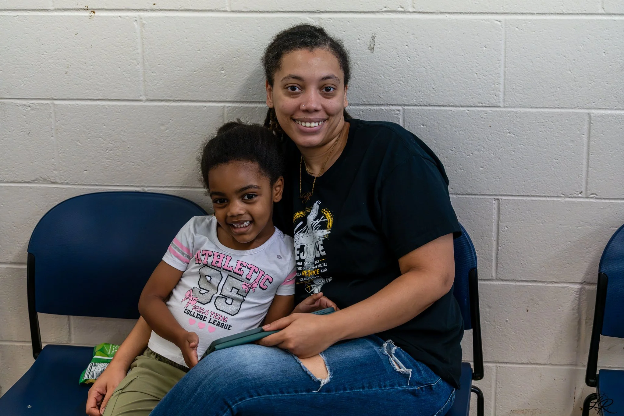 A young girl and a woman sitting together on blue chairs against a beige brick wall, both smiling at the camera. The girl is holding a tablet, and a snack bag is on her lap. The woman has curly hair tied back, a nose piercing, and is wearing a black 