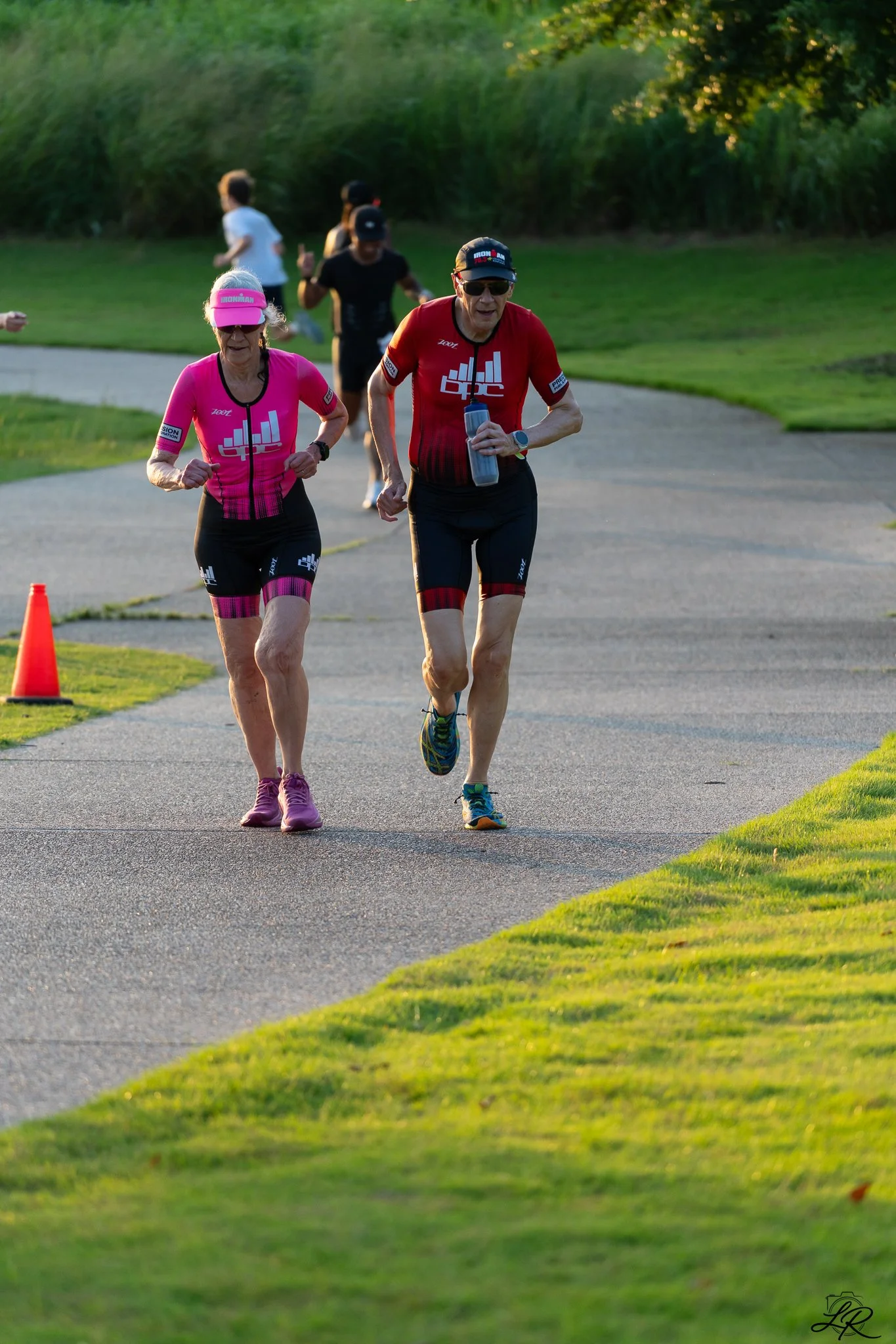Two elderly women running on a paved path, one in a pink athletic outfit with an IRONMAN cap and visor, and the other in a red athletic outfit with an IRONMAN cap, while several people run in the background on a sunny day in a park.