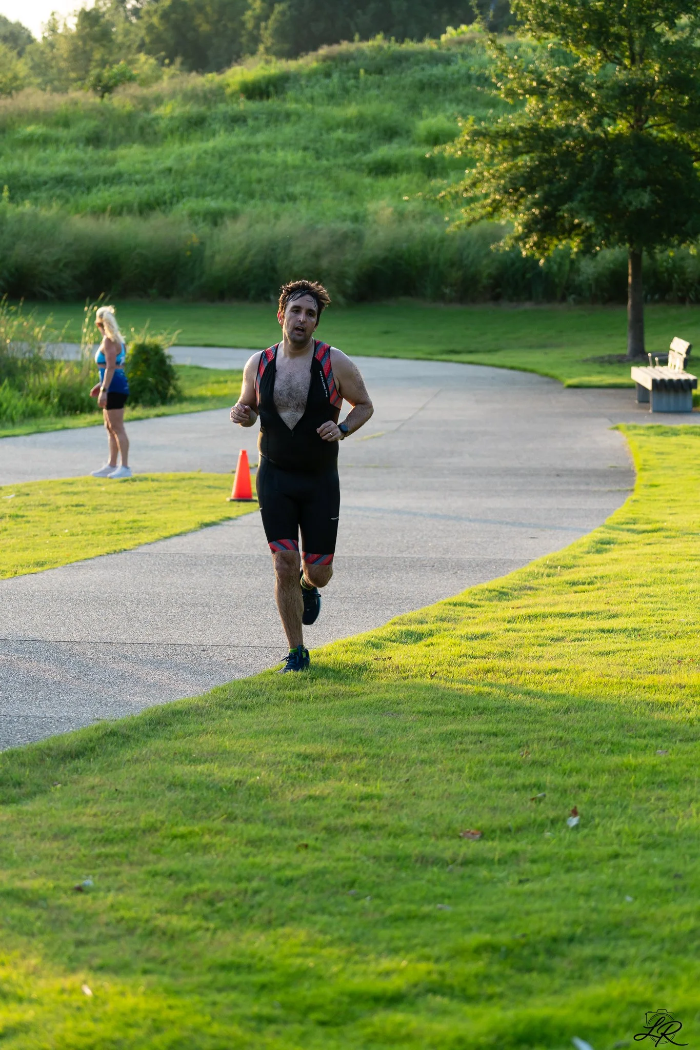 A man running outdoors on a paved pathway in a park during sunset, with trees and a grassy hill in the background, and a woman standing nearby.