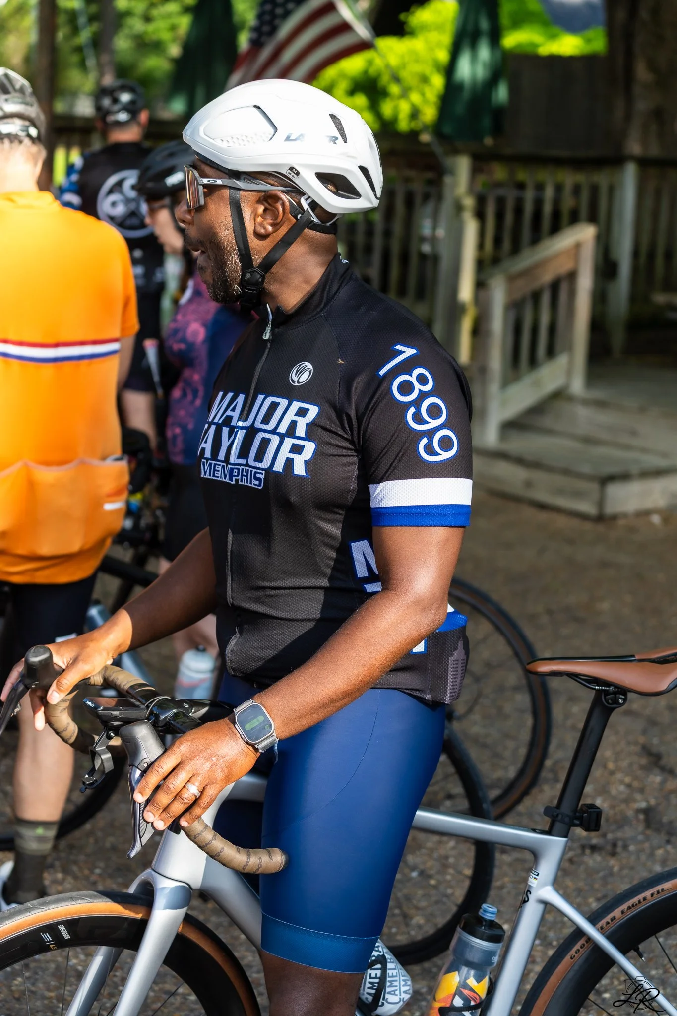 Man wearing a white helmet and black cycling jersey with blue shorts, standing next to a bicycle, surrounded by other cyclists and a wooden fence outdoors.