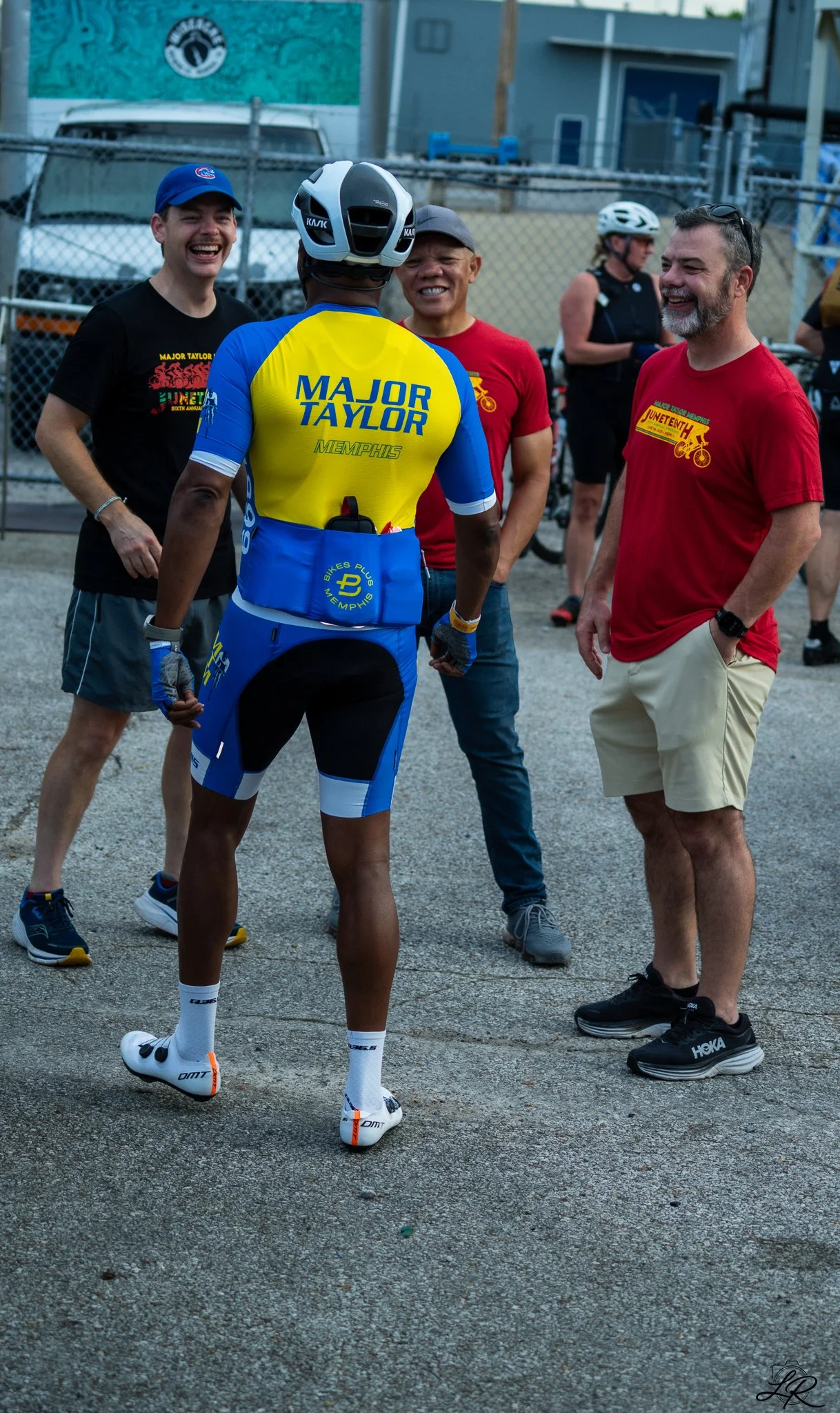 Group of people talking outdoors, one in a cycling uniform and four others in casual attire, all smiling and standing on a gravel surface with a fence and buildings in the background.