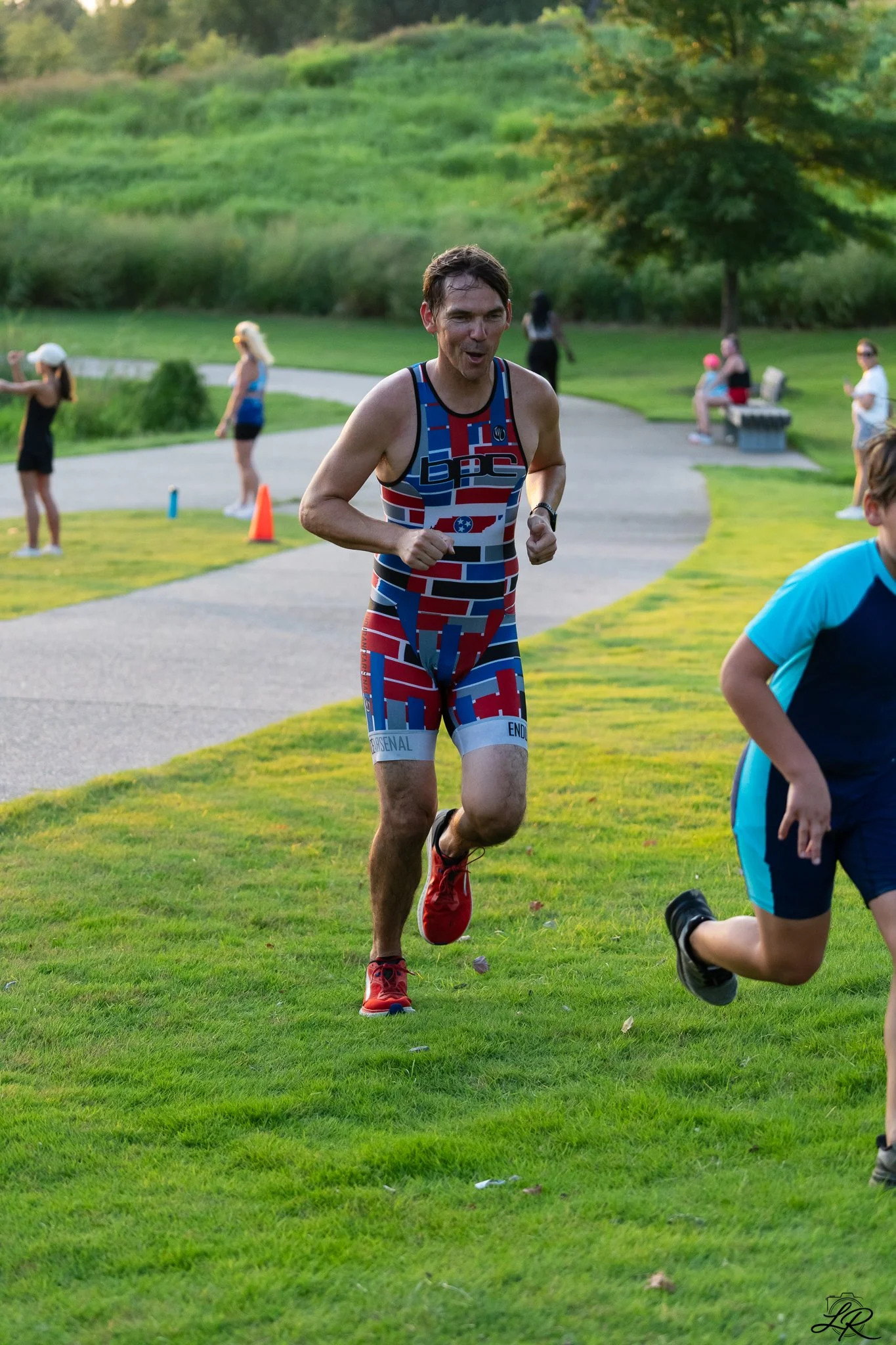 A man participating in a race or running event in a park, wearing a colorful athletic outfit, with a curved pathway and park benches in the background, and several spectators or other participants watching.