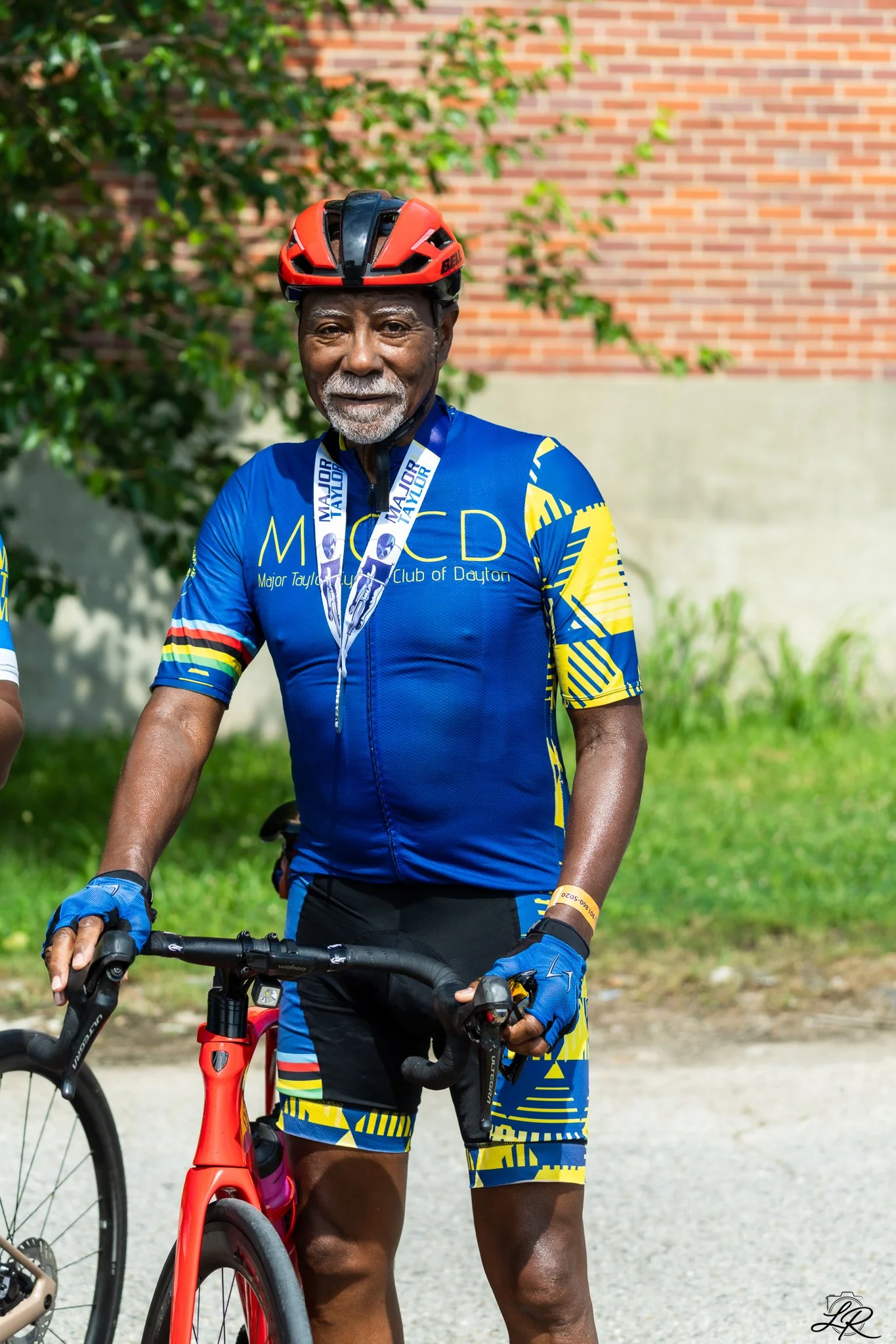 An elderly man wearing a blue and yellow cycling uniform with a medal around his neck, a red and black bicycle helmet, and cycling gloves, standing outdoors with greenery and a brick wall in the background.