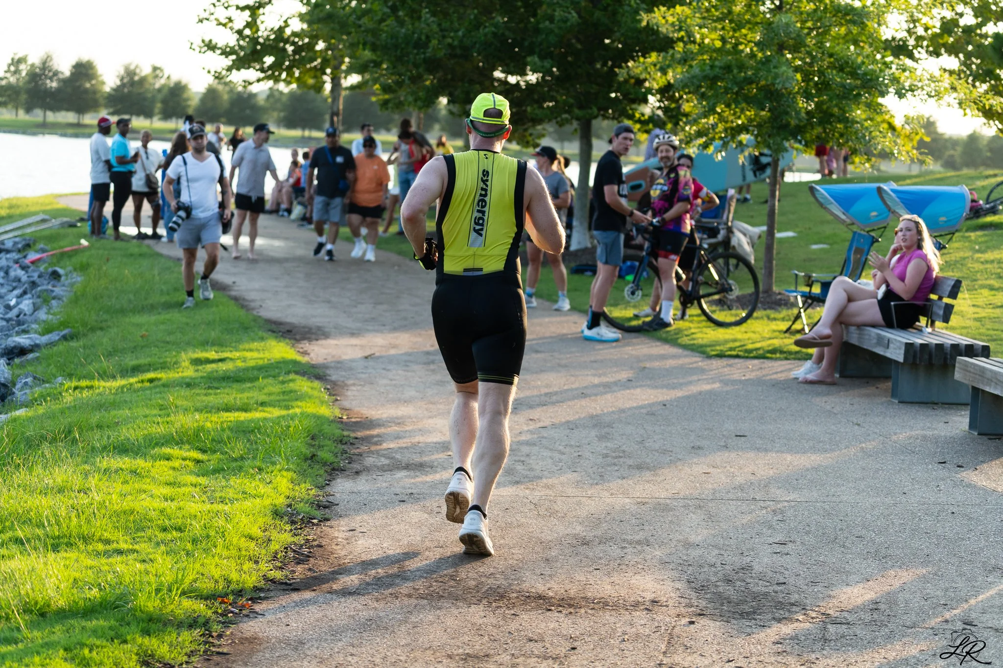 A man in a yellow triathlon suit running on a path by a lake during a race, surrounded by spectators and other participants in a park, with trees and green grass in the background.