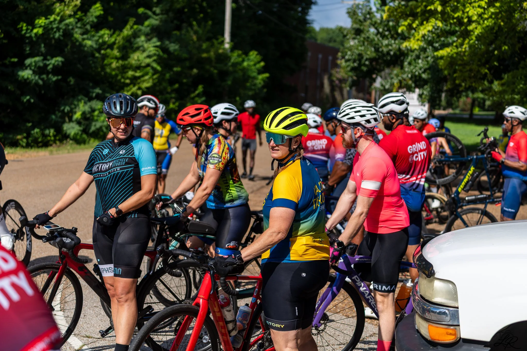 Group of cyclists in colorful jerseys and helmets gathered outdoors on a sunny day with lush green trees in the background.