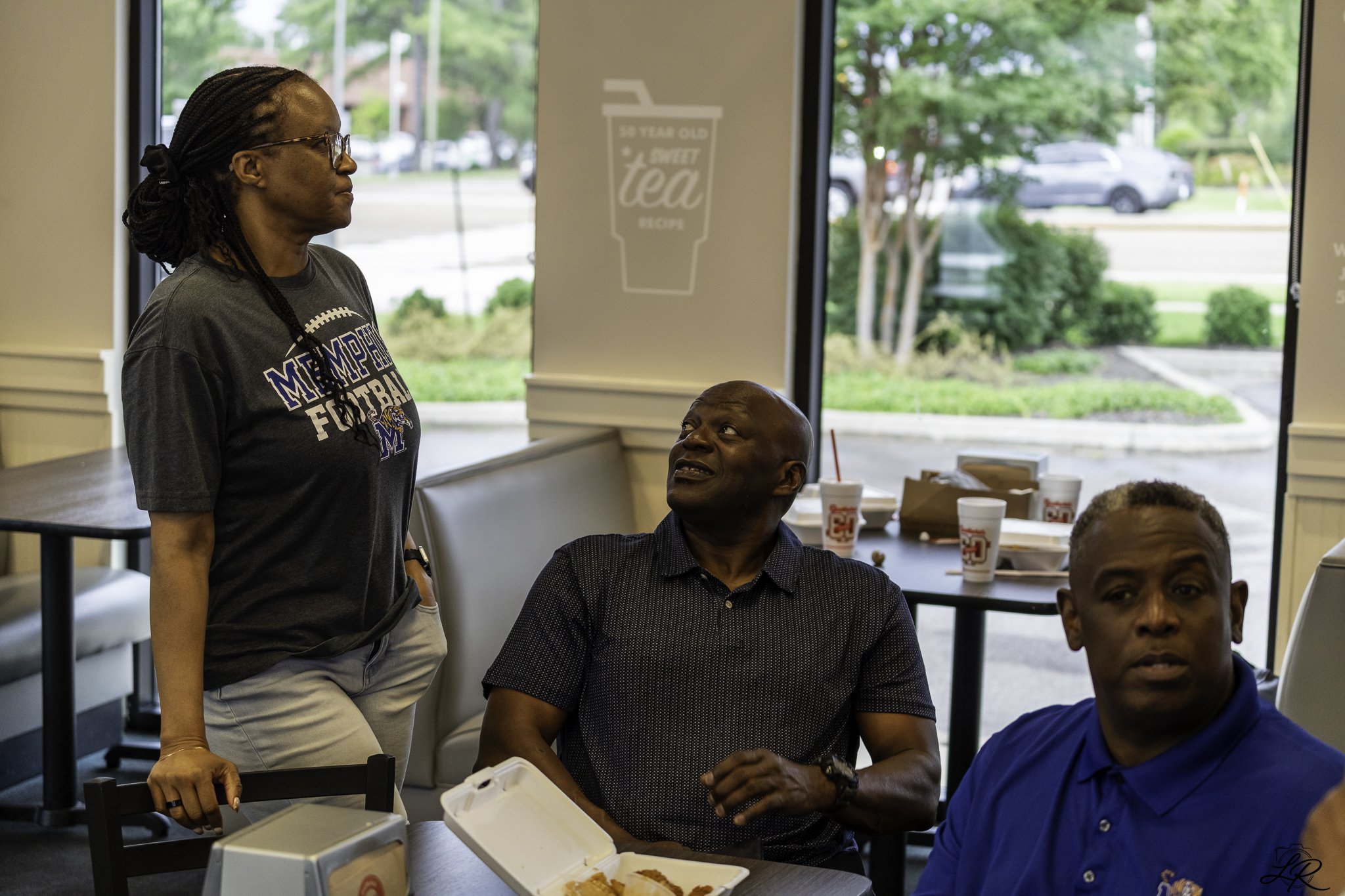 A woman wearing glasses and a Memphis football shirt stands inside a restaurant near a table, while an older man in a dark shirt sits and looks up at her. Several fast-food drinks and food containers are on the table, with large windows in the backgr