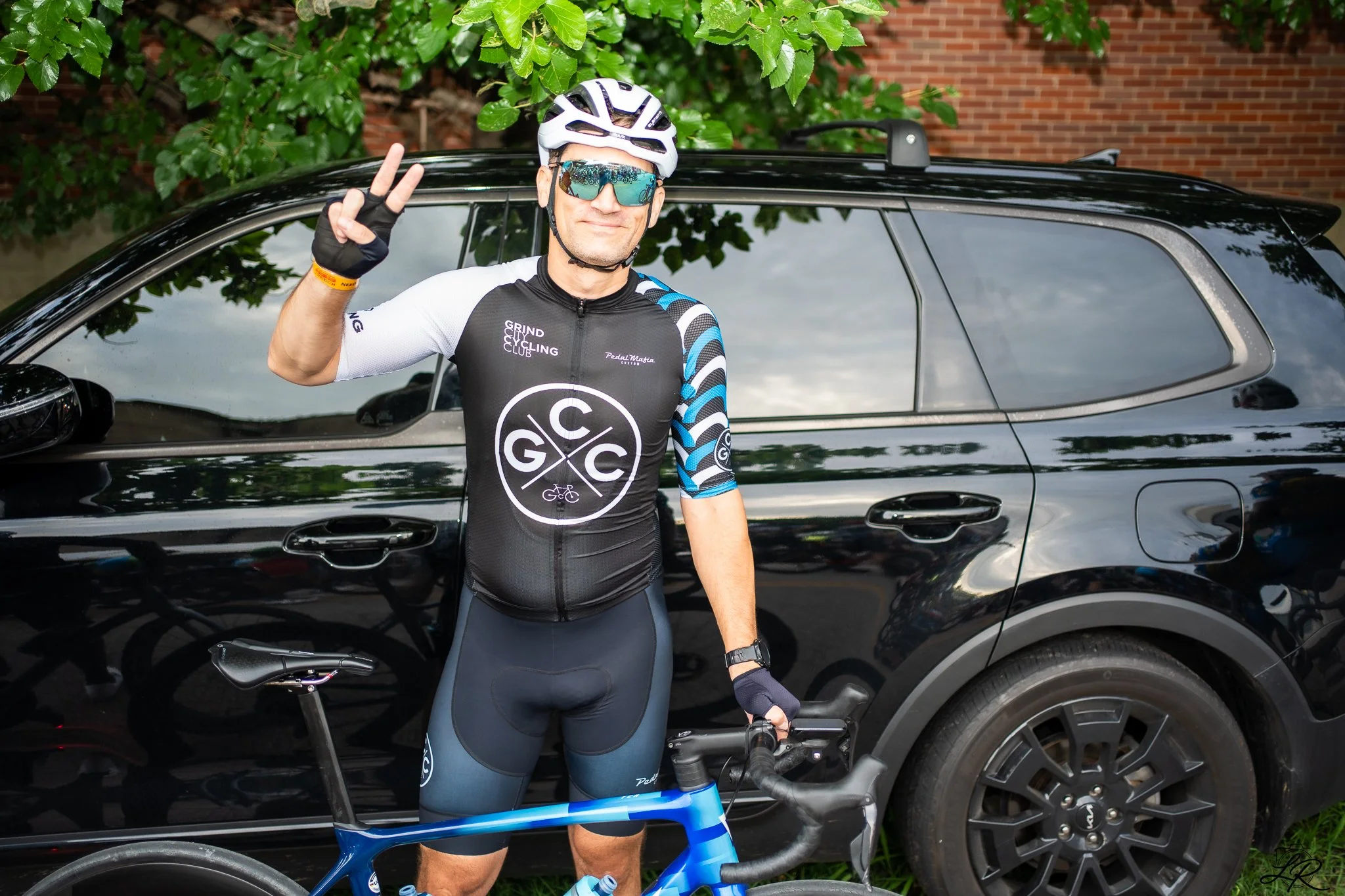A man in cycling gear, including a helmet and sunglasses, standing next to his bicycle with one hand raised making a peace sign in front of a black car and green foliage with a brick building in the background.