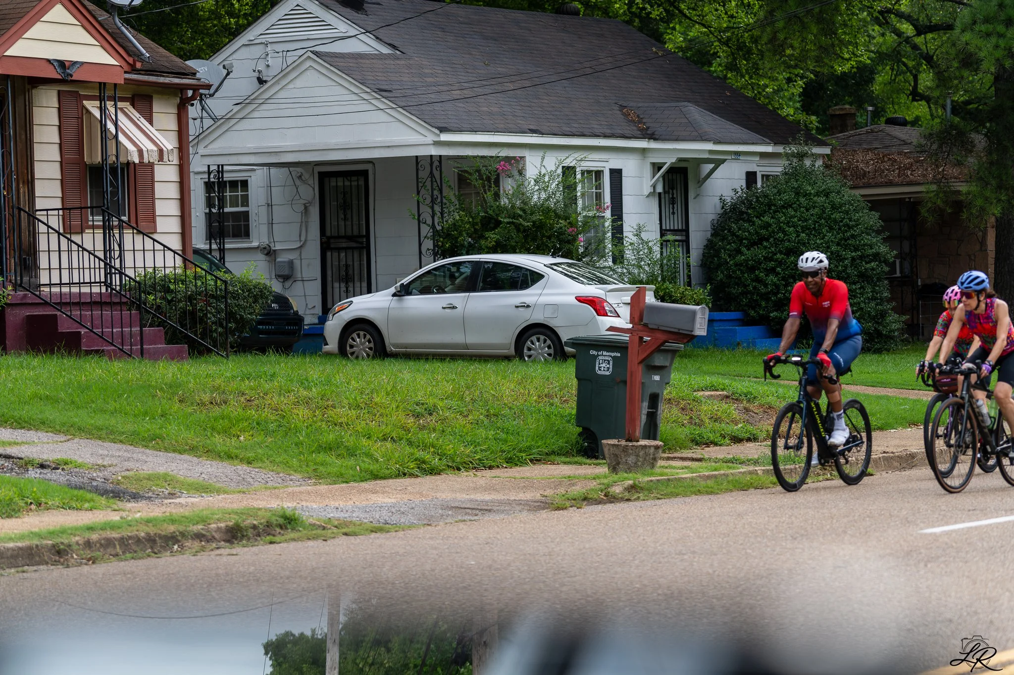 A suburban street scene featuring two houses, one with beige and burgundy accents and the other white, with cars parked in front. There are two children and an adult riding bicycles along the sidewalk, and a green trash bin labeled 'City of Memphis' 