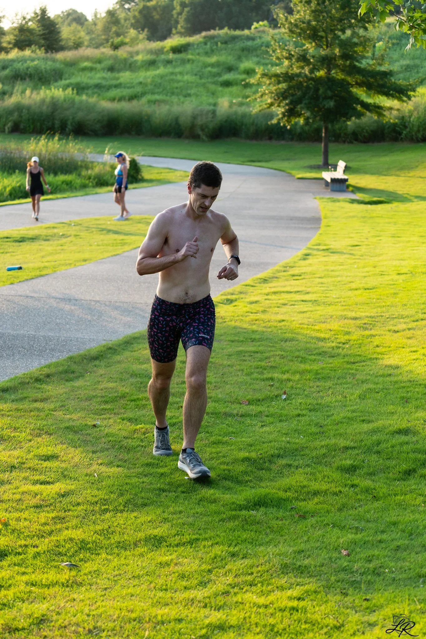 A man running shirtless on a grassy park pathway during sunset with three women walking in the background and a park bench nearby.