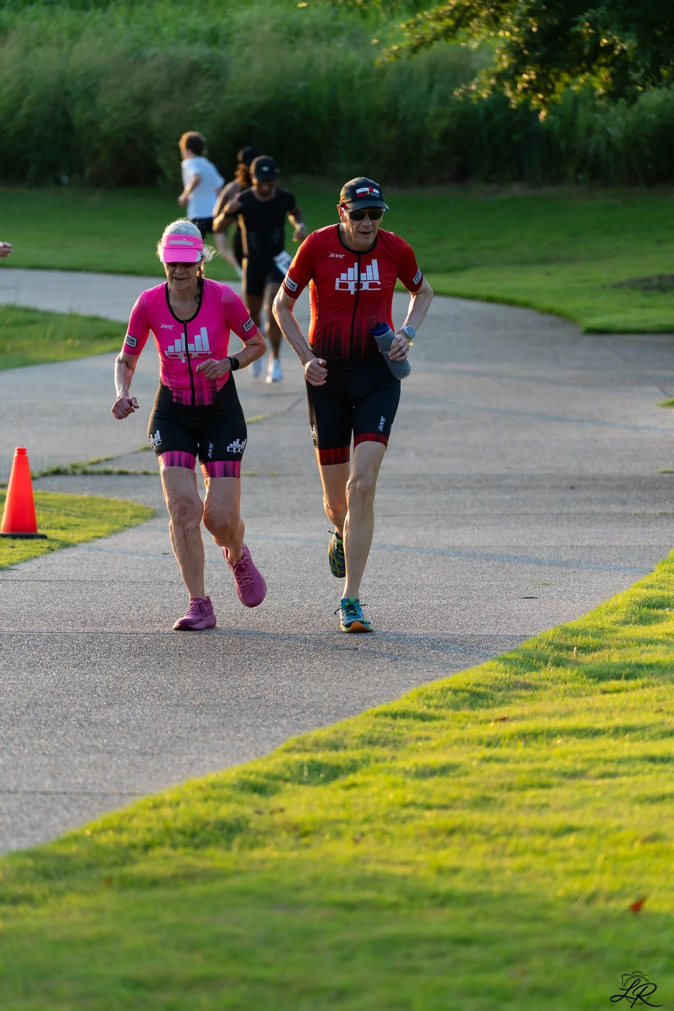 Two older adults running on a paved trail in a park during the evening. The woman is wearing a pink athletic outfit with a visor, and the man is wearing a red and black athletic outfit with a cap and sunglasses. There are other people jogging in the 