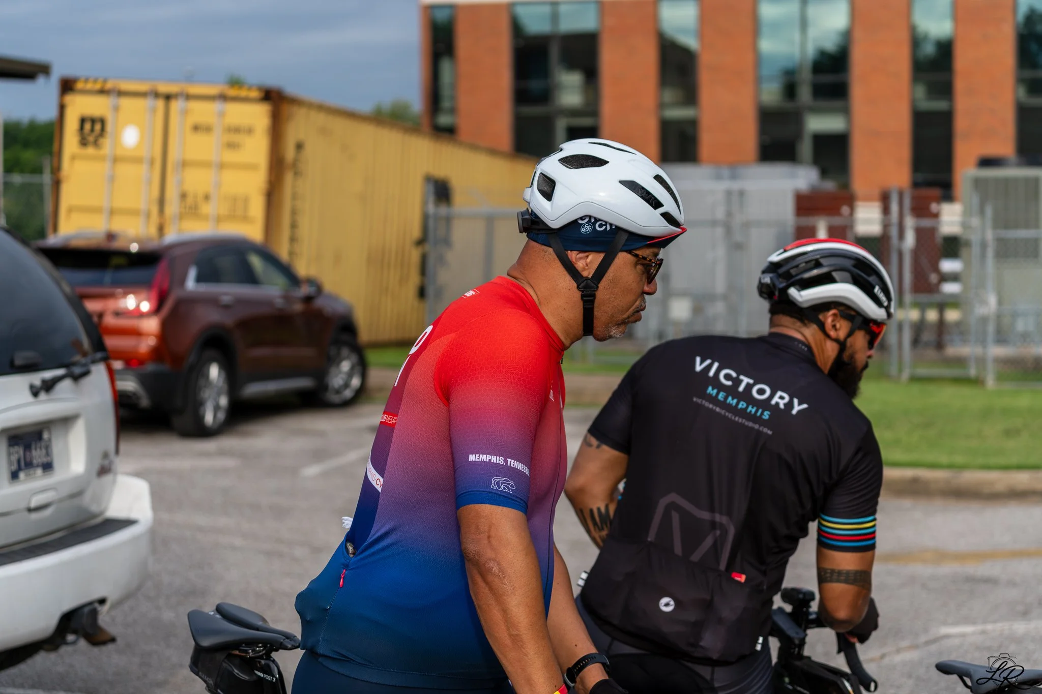 Two men wearing cycling helmets and sports shirts, one in red and blue and the other in black with colorful wristbands, gather near parked cars in an urban area with trucks and a brick building in the background.