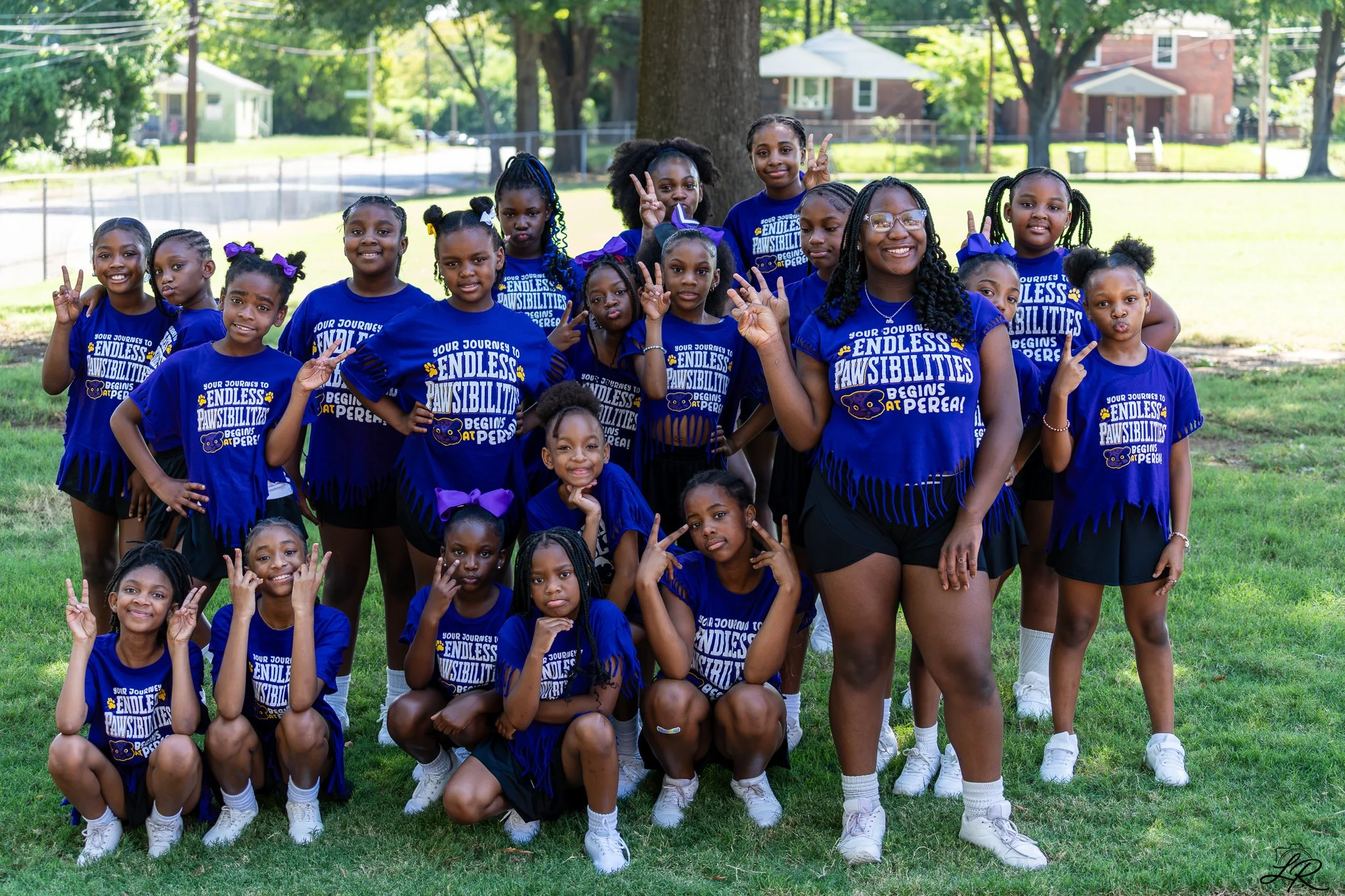 Group of young girls and a woman, all wearing matching blue T-shirts that say 'Your journey to endless pawabilities begins at P.E.R.E.A.', posing outdoors in a park with trees and houses in the background. Most of the girls are smiling and making pea