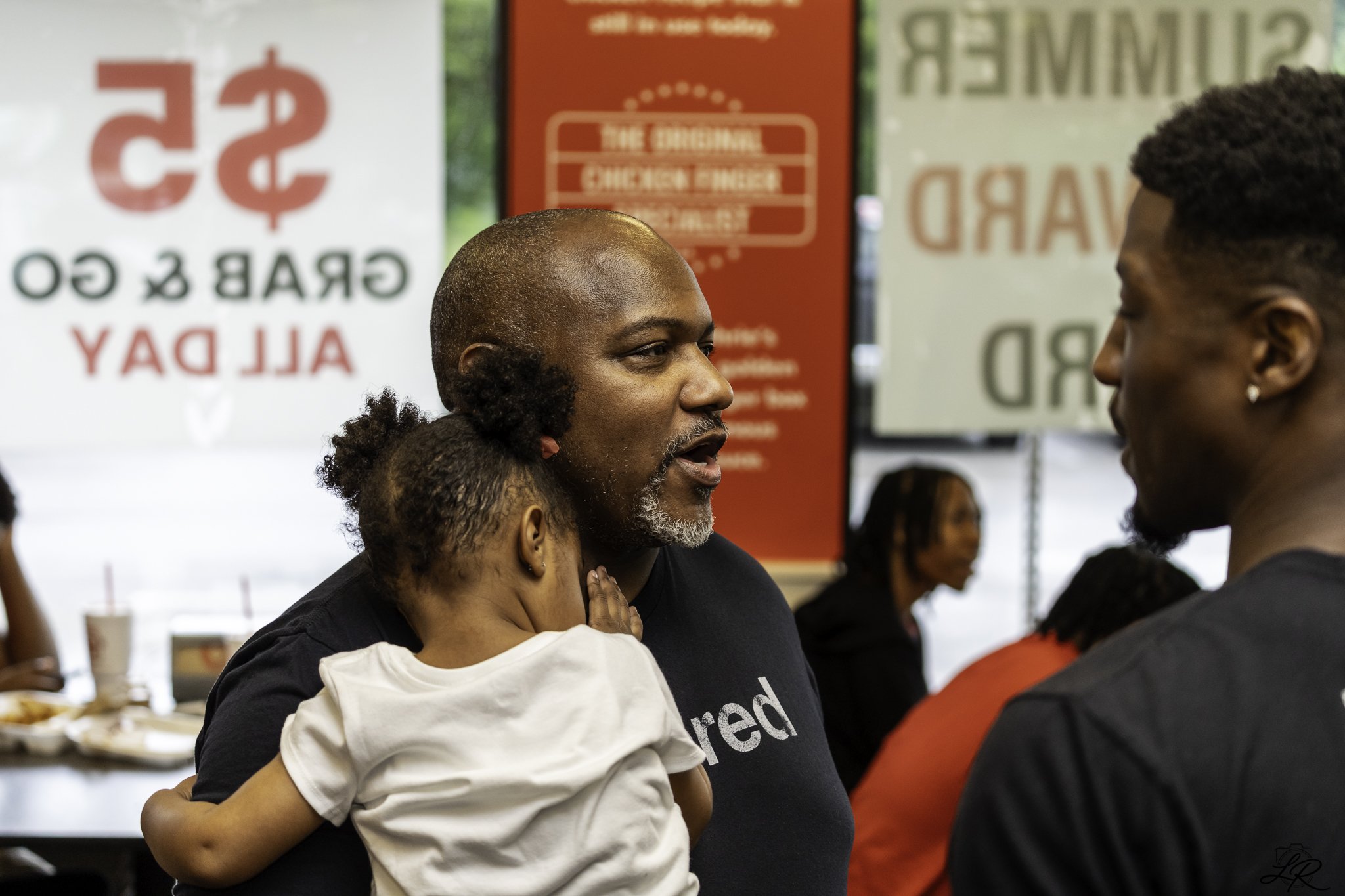 An African American man with a gray beard and shaved head is holding a young girl with curly hair in his arms. They are engaged in a close conversation in a busy setting, possibly a restaurant or food event, with a red sign and people in the backgrou