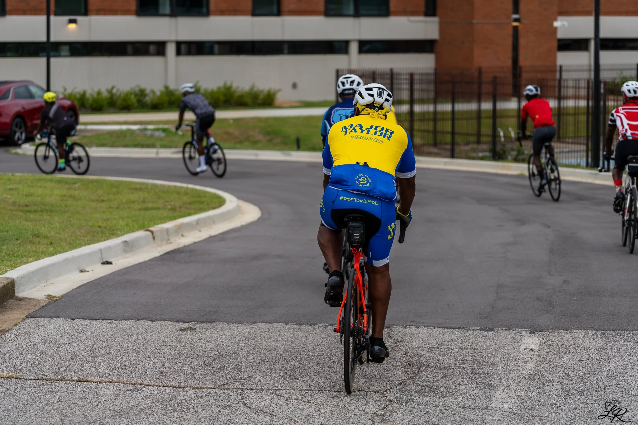 A group of cyclists riding on a paved road near a building, with one cyclist in a yellow and blue jersey in the foreground and others in various colorful outfits in the background.
